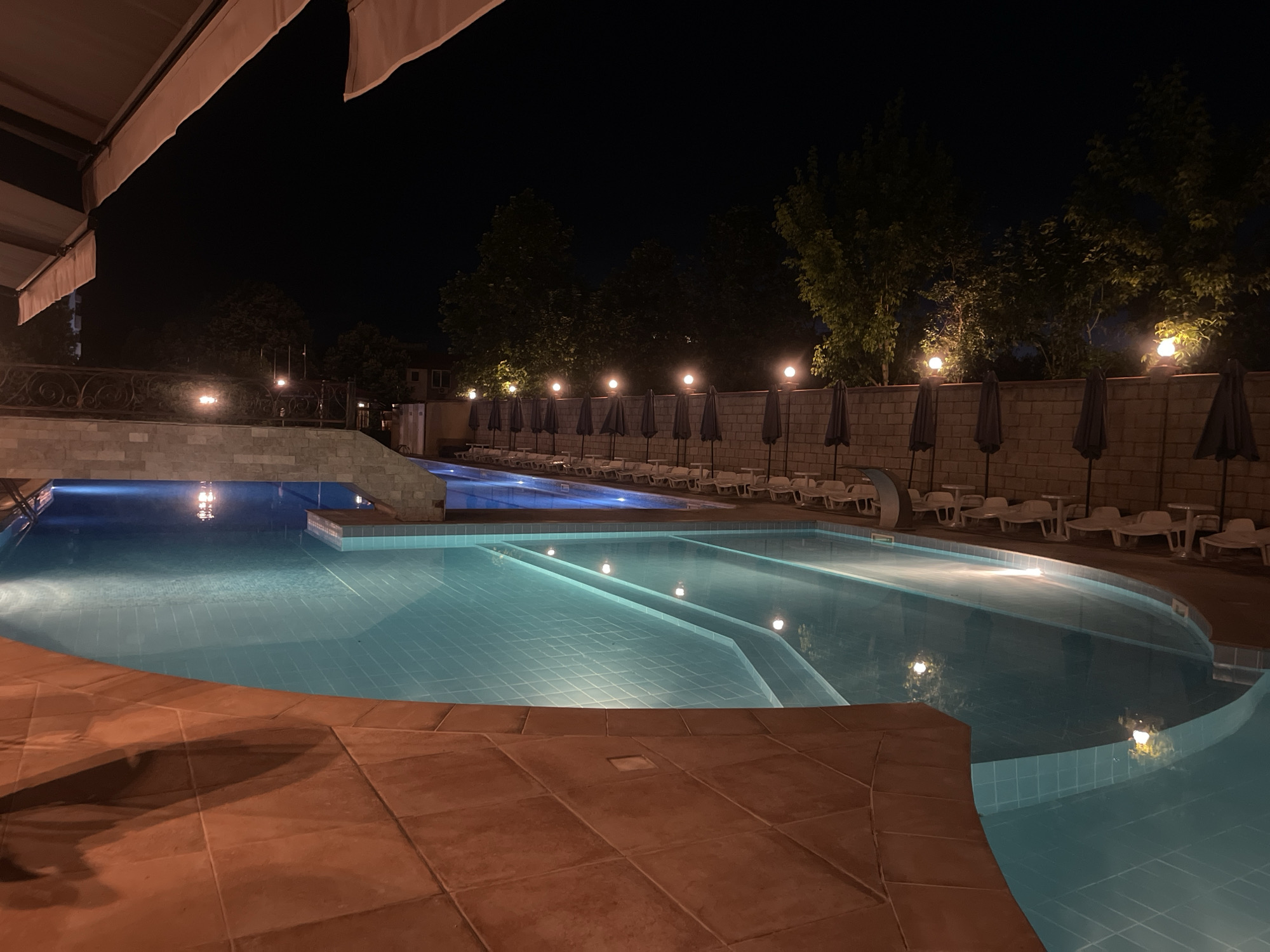 The image shows an outdoor swimming pool area at night. The pool is illuminated from within, casting a blue glow on the water. The surrounding area is paved with terracotta tiles. In the background, lounge chairs and umbrellas are neatly arranged along the poolside. A brick wall with lights and trees behind it encloses the area. The overall scene suggests a resort or hotel setting in Ruse, Bulgaria.