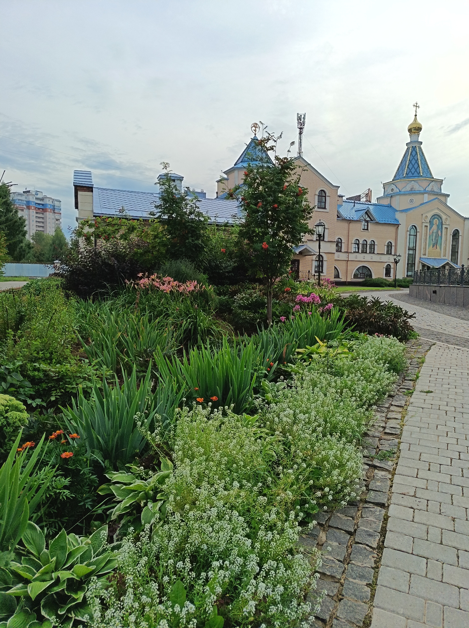 The image depicts a view of the Church of the Holy Trinity in Izhevsk, Russia. The foreground is dominated by a lush, well-maintained garden featuring a variety of green plants, including hostas, irises, and smaller flowering plants with white blossoms. Several bright orange flowers are interspersed within the foliage. The garden is bordered by a cobblestone pathway that curves to the right. Behind the garden, the Church of the Holy Trinity is visible, characterized by its light-colored facade, blue tiled roofs, and a golden dome topped with a cross. To the left of the church, a tall, modern apartment building with a blue and white exterior stands against a cloudy sky. The overall scene suggests an urban environment with a focus on religious architecture and landscaping.
