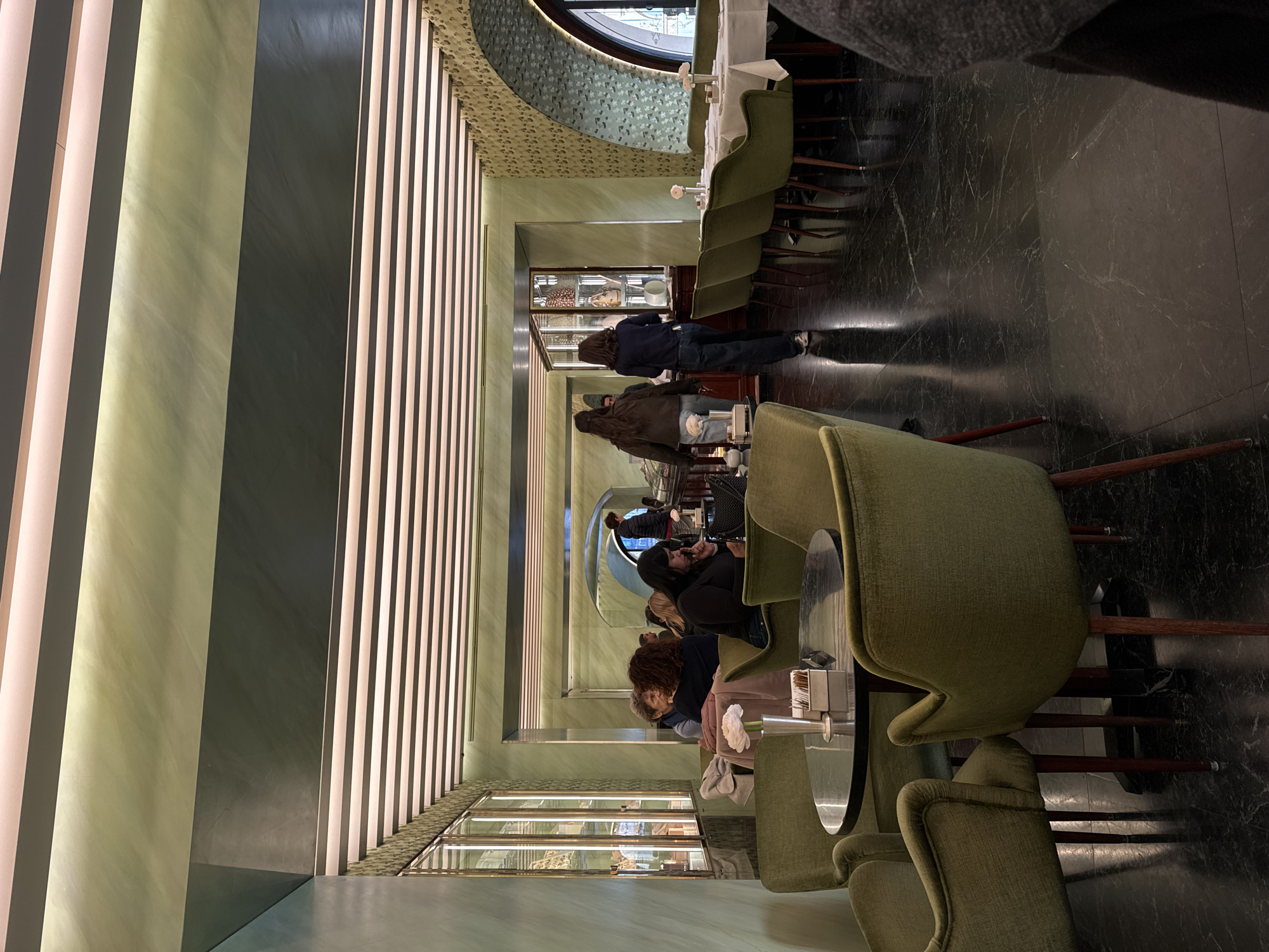 The image depicts the interior of Bar Luce at Fondazione Prada in Milan, Italy, captured from a slightly elevated perspective. The setting is a café or bar characterized by its distinctive decor.
The environment features light green, possibly marble or polished stucco, walls with prominent veining. Sections of the walls, particularly on the right side and near the back, are adorned with a patterned wallpaper in shades of green and beige. The ceiling is striped with alternating dark bands and illuminated light panels running horizontally. The flooring consists of large, polished dark marble tiles with subtle lighter veining.
In the immediate foreground, a partial view of a person's right shoulder and arm, clad in dark fabric, is visible on the far right. To the left of this, a round, dark-topped table is surrounded by four olive-green velvet upholstered chairs with dark wooden legs. On this table, a silver cylindrical vase holds a single white flower, and a white rectangular dispenser box for sugar or sweeteners is present.
Further into the mid-ground on the left, several individuals are seated. A woman with dark, wavy hair is observed from behind, facing away. To her right, another woman with dark hair looks down at a smartphone in her hands. Next to her, an older woman with short grey hair is visible from the side, appearing to engage in conversation. Along the left wall, multiple tall, glass-fronted display cases with metallic frames showcase various items.
In the center-right mid-ground, two women are walking away from the camera, moving towards a counter or bar area in the background. The leading woman wears a dark jacket and light-colored jeans, while the one behind her wears a dark blue top and dark jeans. The counter area appears to have a person behind it and display cases with various objects.
On the right side of the mid-ground, a row of dining tables draped with white tablecloths is arranged. Each table features a small vase with a light pink flower. These tables are accompanied by the same olive-green velvet chairs. An arched window, partially visible on the far right, suggests a view to the exterior. The overall lighting is predominantly artificial, emanating from the ceiling strips, creating a warm ambiance.