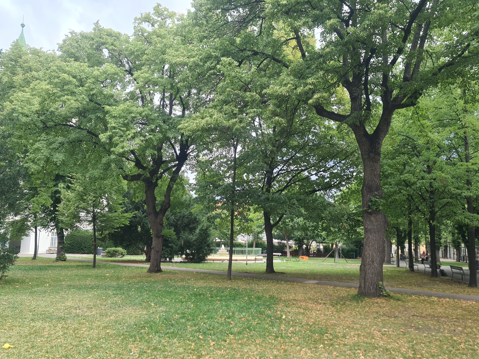 The image depicts a park scene in Vienna, Austria, likely on a cloudy day given the overcast sky. In the foreground, a grassy lawn is sparsely covered with fallen leaves. A paved pathway curves through the park, with several benches visible along its edge. Large, mature trees with lush green foliage dominate the landscape, casting shadows on the grass. To the left, a white building with arched windows and a green spire is partially visible behind the trees, suggesting a historic structure. In the background, there is a playground area with a green fence and some play equipment. People are seen sitting on benches and walking in the distance, indicating public use of the park. The overall impression is one of a peaceful, urban green space.