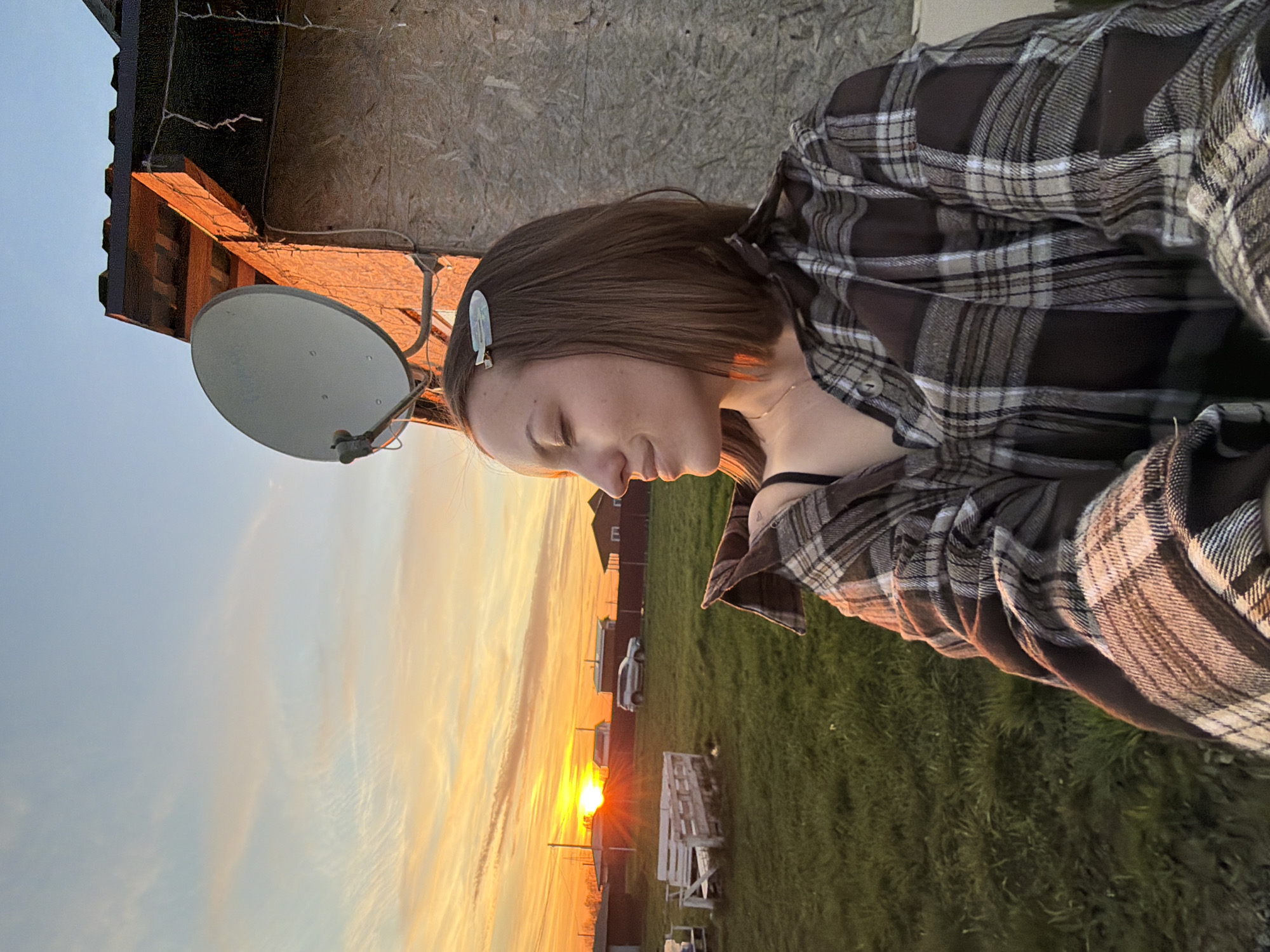 A medium shot captures a woman wearing a brown plaid shirt against a backdrop of a rural sunset in Razdol'ye, Russia. The woman has short brown hair and a decorative clip. A satellite dish is mounted on a building behind her. The sunset illuminates a field with scattered objects and distant buildings. A car is parked nearby.