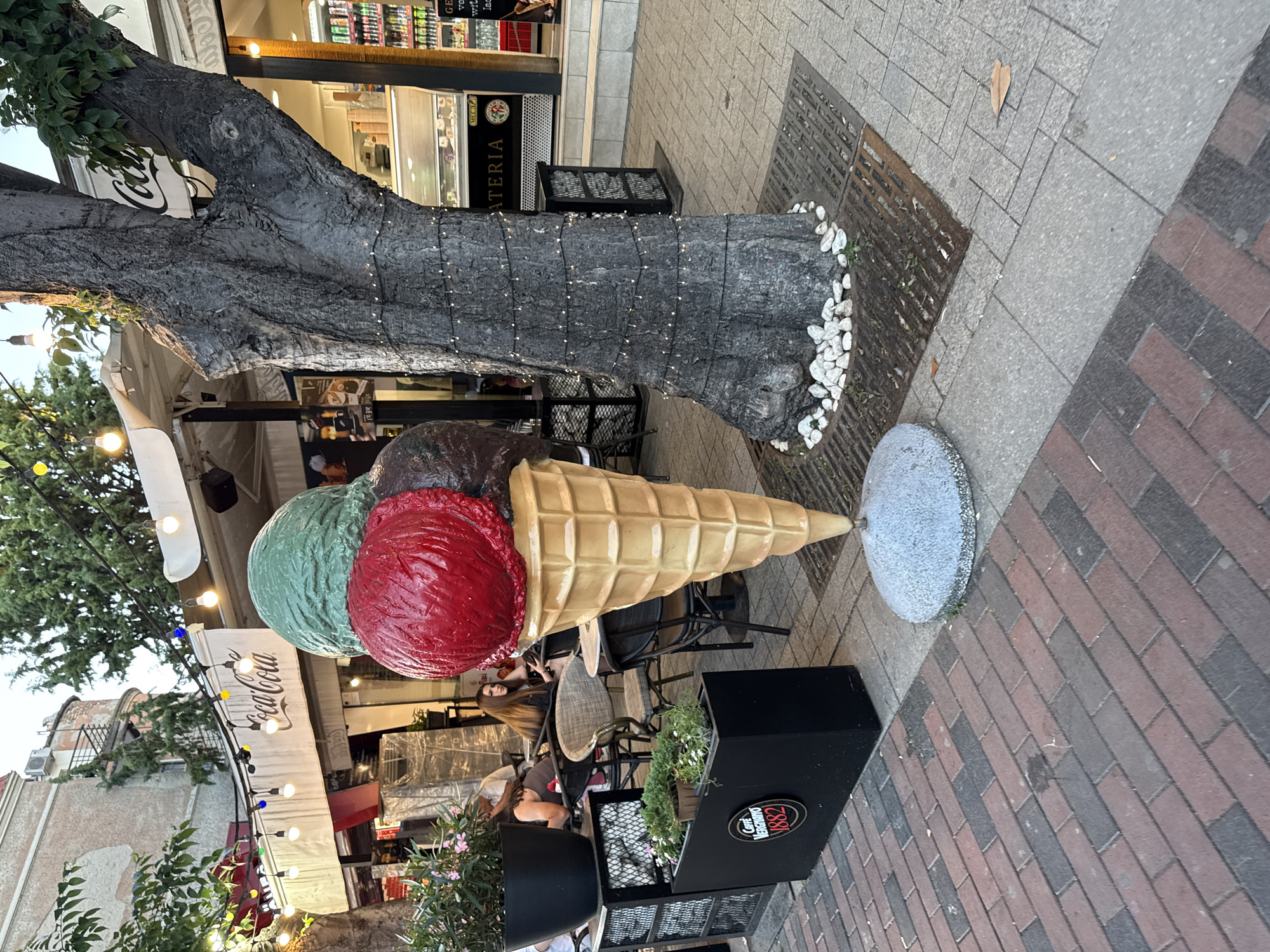 The image depicts an outdoor scene in an urban environment. A large, three-scoop ice cream cone sculpture is prominently displayed in the foreground, with scoops of green, red, and brown ice cream. It is positioned on a gray concrete base. To the right of the sculpture is a large tree with a thick trunk, adorned with strings of small white lights. The tree's base is surrounded by small white pebbles. The ground is paved with a mix of brick and concrete.
In the background, there is a building with a white awning and string lights overhead. A sign with "Coca-Cola" is visible on the awning. Below the awning, there are tables and chairs set up, suggesting an outdoor seating area for a cafe or restaurant. Several people are seated at the tables. A black planter box with green plants is to the left of the ice cream sculpture. Further in the background, a shop window displays merchandise, and a refrigerator unit is visible. The overall atmosphere suggests a pleasant daytime scene, possibly in a pedestrian-friendly area of a city.