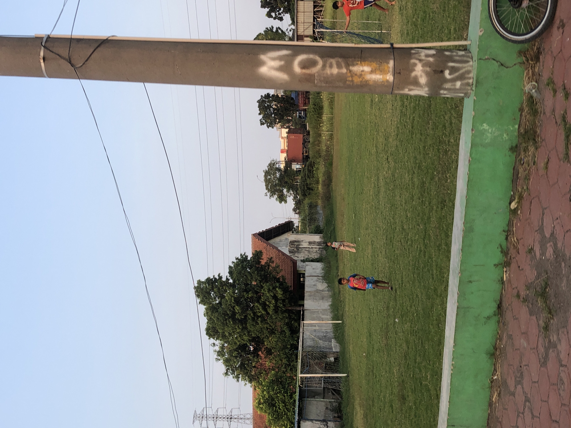 In Wunut, Indonesia, a grassy field serves as a makeshift football pitch where several children are engaged in play. A young boy in a red shirt and blue shorts stands on the field, while another child in beige trousers stands further back. A football goal is visible on the left side of the frame, behind a low concrete wall. Beyond the field, a building with a red-tiled roof and a concrete wall stands, partially obscured by trees. A tall utility pole with graffiti is prominent in the foreground on the right, with electrical wires stretching across the sky. In the background, trucks and industrial buildings can be seen, indicating a mixed urban and rural environment. A bicycle wheel is visible at the bottom right of the image.