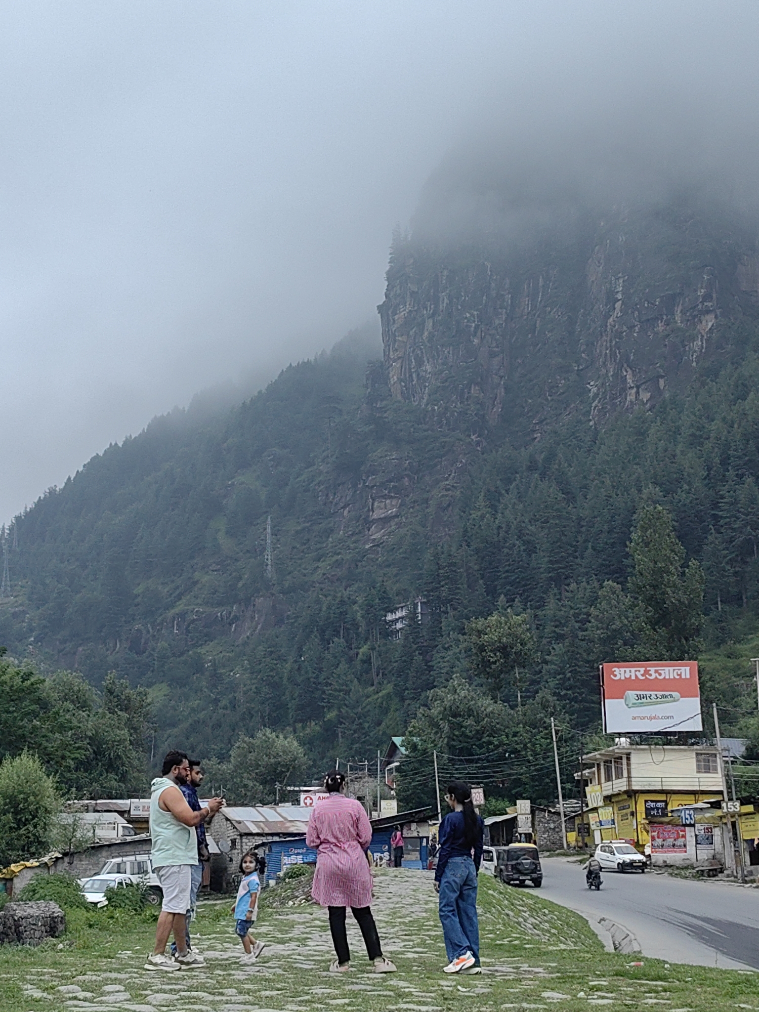 The image captures a scene in the city of Bahang, India, with a towering, fog-shrouded mountain range dominating the background. A dense forest of evergreen trees covers the lower slopes of the mountain. In the foreground, a group of people are standing on a stone-paved path. A man in a light blue sleeveless hoodie and white shorts is taking a photo with his phone. Beside him, another man is also looking at his phone. A young girl in a blue and white dress is walking on the path, looking at the group. A woman in a pink striped top and black pants is facing away from the camera, looking in the same direction as the others. To the right, a road curves through a settlement with buildings and shops. A large red billboard with Hindi text reading "अमर उजाला" (Amar Ujala) is visible on top of one of the buildings. Several vehicles, including cars and a motorcycle, are on the road. The sky is overcast and misty, contributing to the diffused light in the scene.