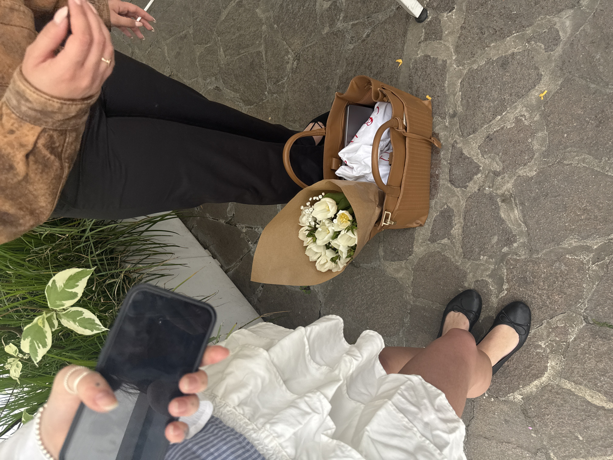 An overhead view captures two individuals seated outdoors on a paved surface, likely in Milan, Italy, given the context. The ground is composed of dark, irregularly shaped cobblestones or pavers with lighter grout lines, bordered by patches of green grass and variegated plants.
One person, positioned on the left, wears a brown jacket, possibly leather, and black trousers. Their right hand, adorned with a ring and light-colored nail polish, holds a thin white object, potentially a cigarette. The second person, at the bottom right of the frame, wears a white, ruffled skirt or dress and black ballet flats. Their bare legs are visible, and their left hand, decorated with multiple rings and a beaded bracelet, holds a black smartphone.
Between the two individuals sits a brown handbag or tote bag on the ground. Inside the bag, a folded white t-shirt with a red design or logo (possibly a heart or text) is visible, along with another dark object. Next to the bag is a bouquet of white flowers, predominantly tulips, wrapped in brown craft paper. Small yellow paper or fabric bits, resembling confetti, are scattered across the paving. A white metal structure, possibly part of outdoor furniture, is partially visible in the upper right corner.
The scene depicts a casual, relaxed outdoor gathering, possibly friends meeting. The presence of the flowers and confetti-like elements might suggest a celebration or a special occasion. The lighting indicates daytime, with no harsh shadows, implying possibly an overcast day or late afternoon. No clear text is legible other than the red design within the handbag.