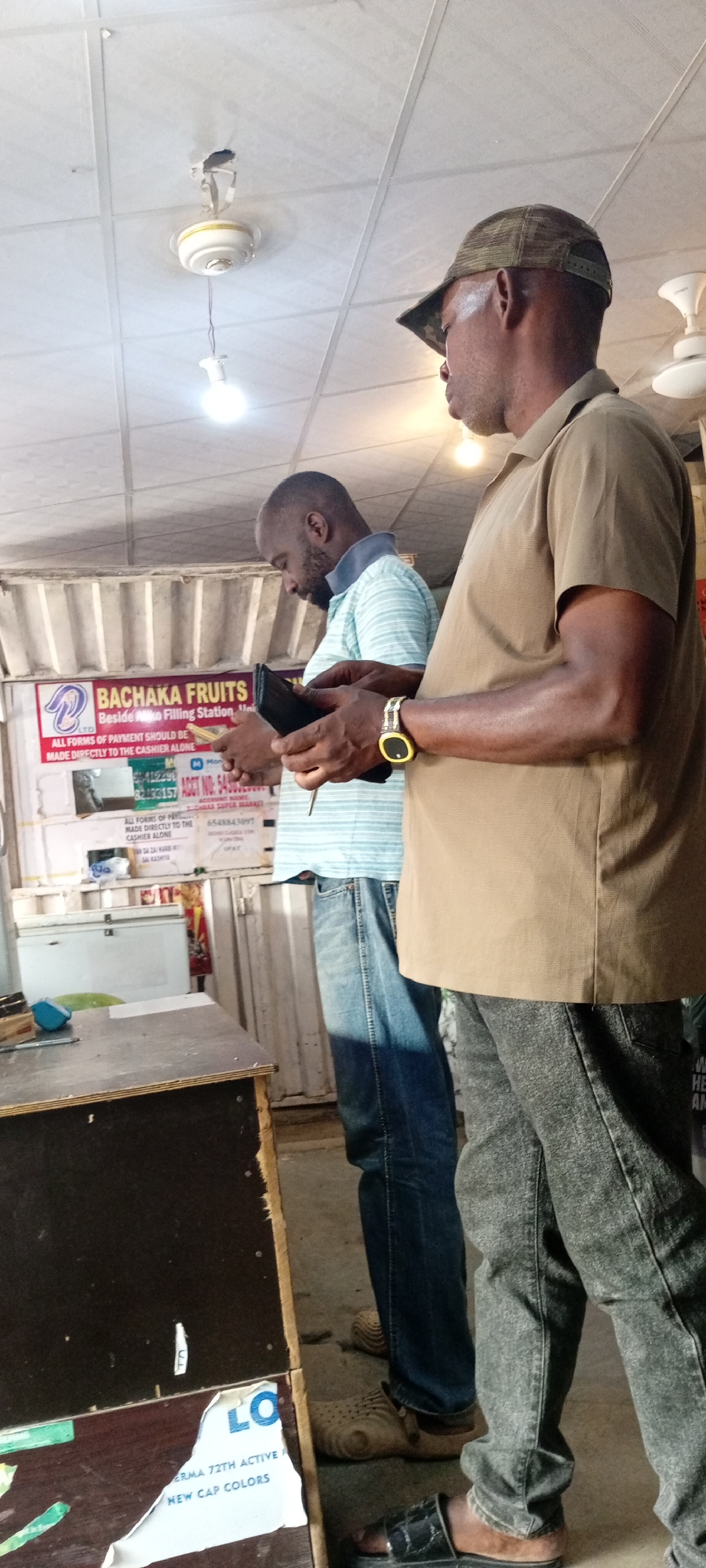 In Aviation Quarters, Nigeria, two men are standing near a stall. The man in the foreground, wearing a camouflage baseball cap and a tan shirt, looks to his right. He has a watch on his left wrist. The man behind him, wearing a light blue and white striped t-shirt and blue jeans, is looking down at his hands, holding a black wallet and a pen. The background shows a stall with a sign that reads "BACHAKA FRUITS" and other informational text. A white cooler is visible behind the stall. The ceiling has a fan and light fixture. The floor is concrete.