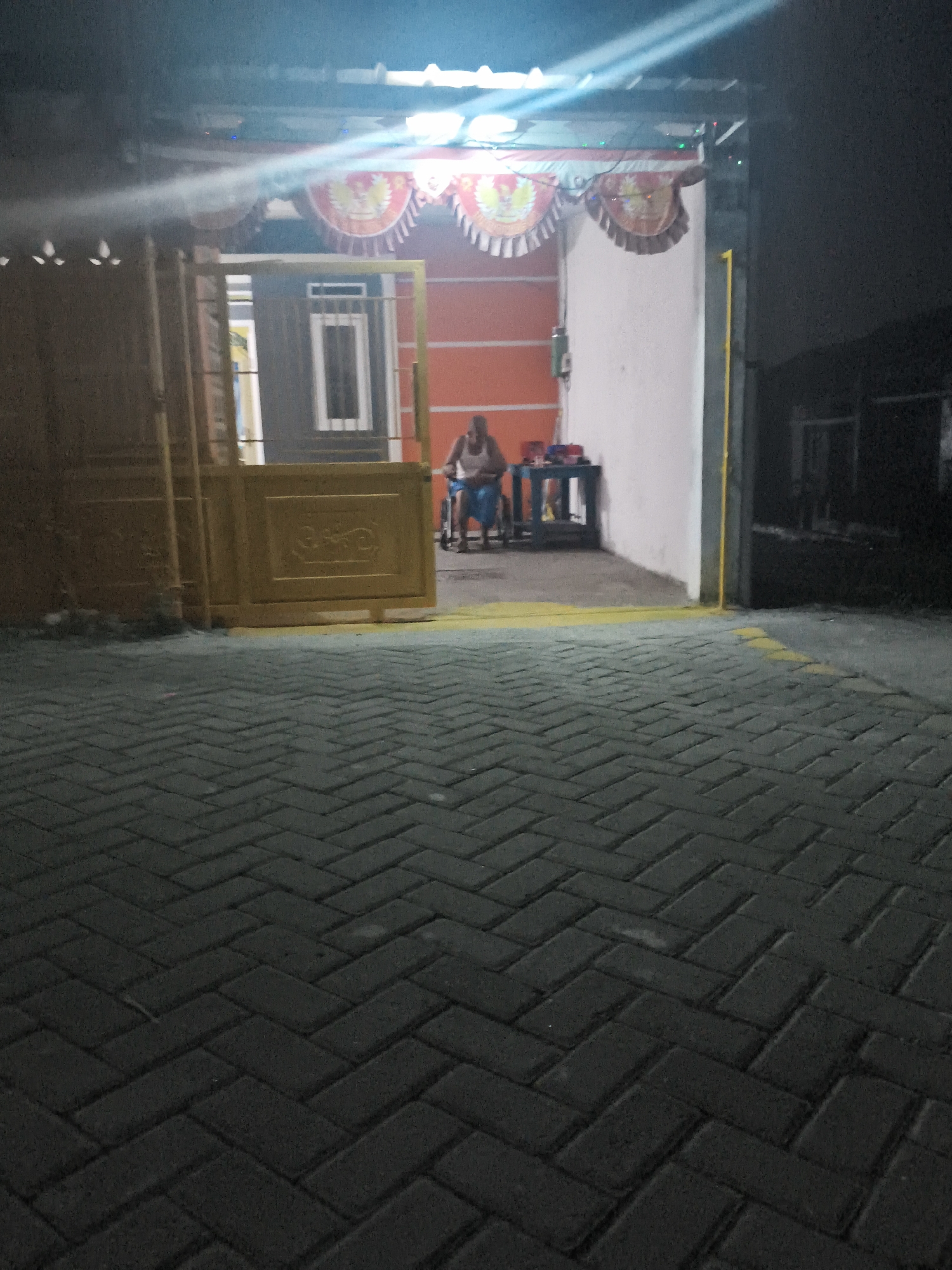 The image depicts a nighttime scene in front of a building, possibly a shop or residence, in Banjar Agung, Indonesia. The foreground is dominated by a dark, herringbone-patterned brick walkway. Beyond the walkway, a yellow gate is partially open, revealing an interior space. Inside, a person is seated in a wheelchair to the right of the gate, facing towards a table with some objects on it. The building's facade is painted in shades of white and orange. Decorative banners with circular emblems are hung above the entrance. Lighting, including bright beams, illuminates the scene from above and within the building, creating a slightly hazy atmosphere. The overall impression is one of a quiet evening in a semi-urban Indonesian setting.