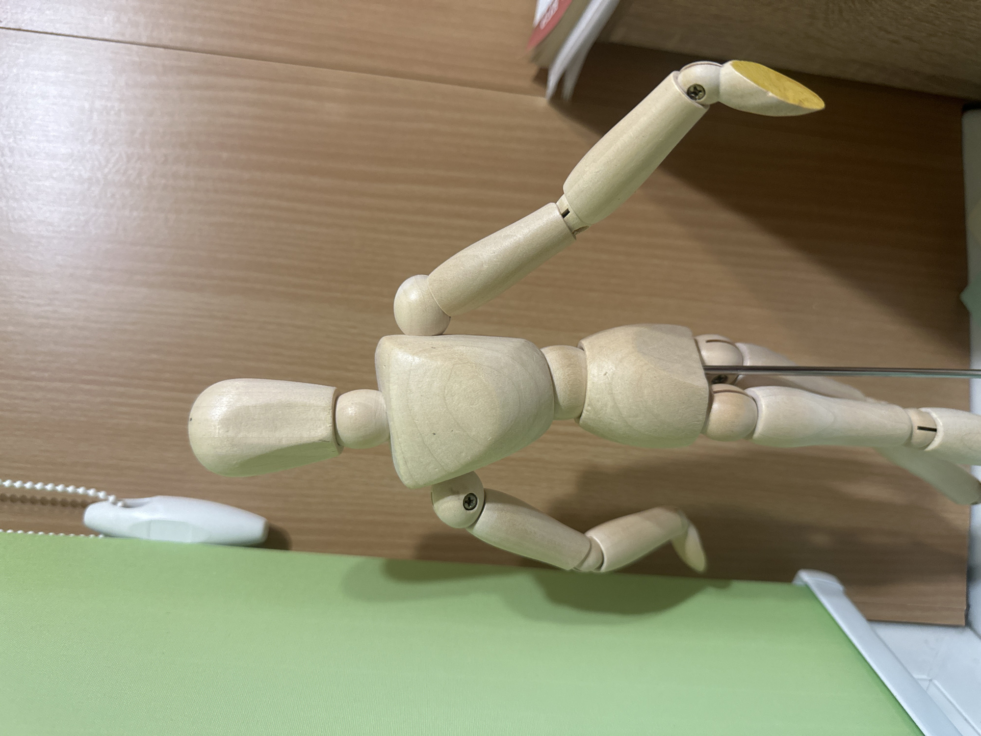 A wooden artist's mannequin is depicted horizontally, posed dynamically as if running, leaping, or flying. The mannequin, crafted from light-colored wood with visible joint mechanisms and screws, is suspended in mid-air by a slender metal rod that enters its back. The figure's left foot features a distinct yellow tip.
The scene is set indoors, with the mannequin positioned against a backdrop of a light brown wooden surface, possibly a desk or shelf, exhibiting a visible wood grain. Below the mannequin and to its left, a vibrant lime-green surface, likely a mat or a large book, occupies the foreground. Resting on this green surface, near the mannequin's head, is a white cord pull with a beaded chain. The lighting is even and soft, suggesting an indoor environment without specific time-of-day indicators. There is no visible text within the image. While the image itself contains no explicit location cues, the provided context places this scene in Kazan, Russia.
