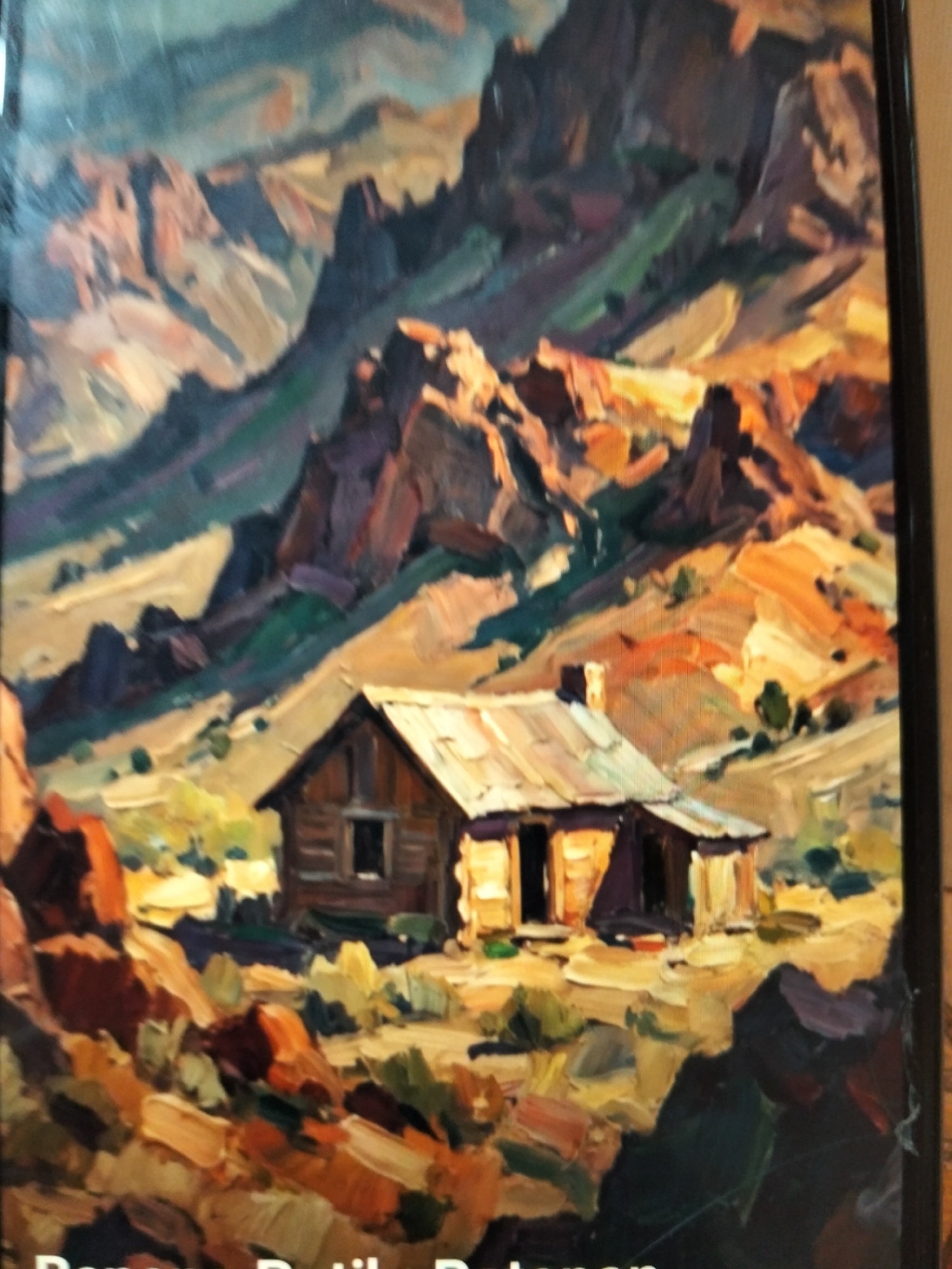 The image is a painting of a rugged, mountainous landscape with a small wooden cabin in the foreground. The mountains are depicted with bold brushstrokes in shades of purple, brown, and yellow, suggesting a dry, arid environment. Sunlight casts strong shadows, highlighting the textures of the rock formations. The cabin is simple, with a tin roof and a dark doorway. Small green shrubs are scattered around the cabin and in the middle ground. The painting style is impressionistic, with visible texture and a focus on light and color. The bottom edge of the image shows part of a black frame. The text at the very bottom of the image is not legible. The location is not identifiable from the image.