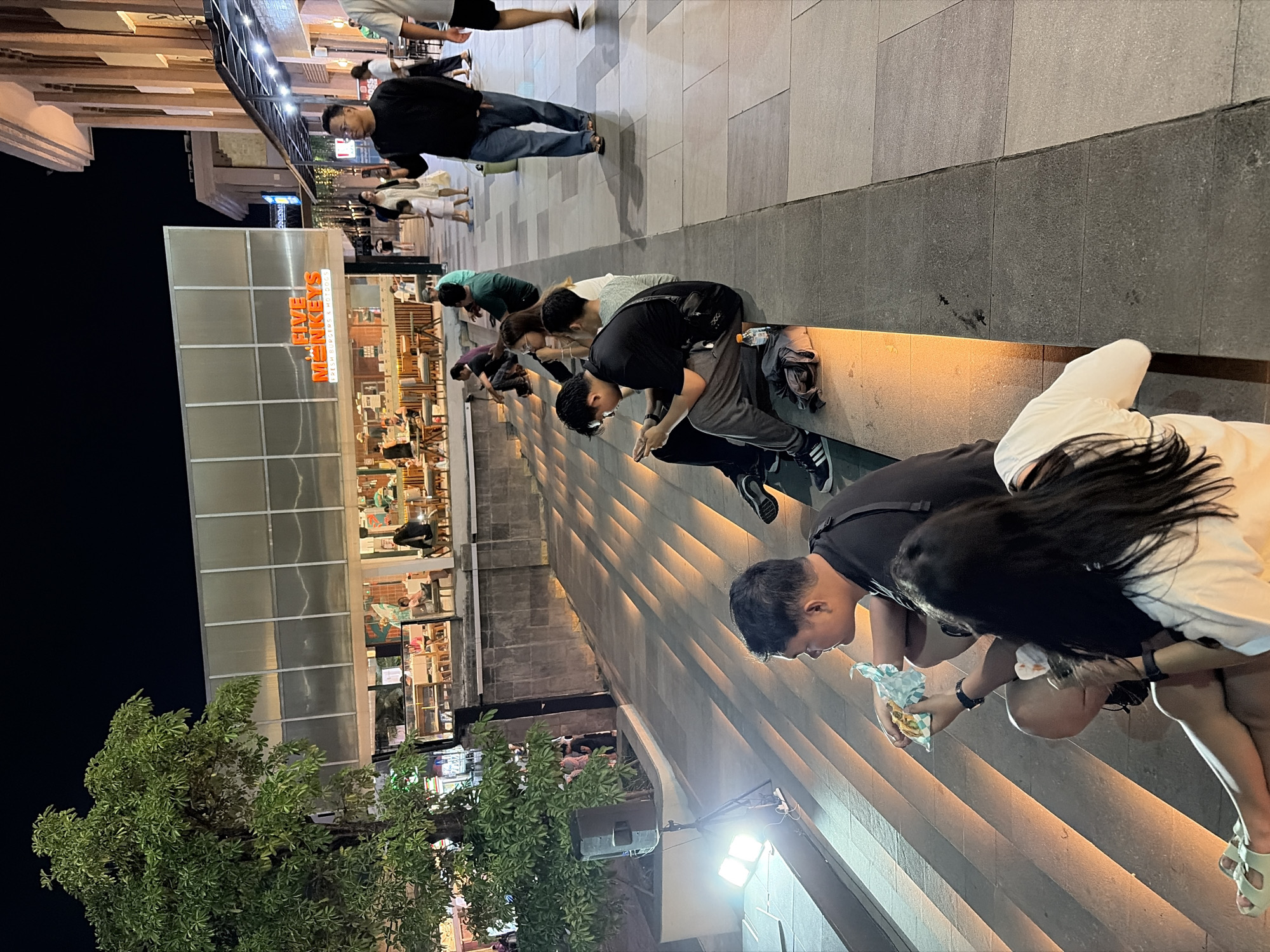 A group of people are seen on a concrete staircase outdoors at night. The lighting suggests it is evening or nighttime, with artificial lights illuminating the scene. The individuals are dressed casually, wearing t-shirts, shorts, and pants. Some are standing, while others are seated or in a crouching position on the stairs.
One person in the foreground, wearing a white top and black pants, is holding a burger and appears to be eating it. Next to them, another person in a black top is also looking down, possibly engaged in eating or using their phone. A group of people further up the stairs are in a similar crouching posture, with their heads bowed. The setting appears to be an urban environment, with a building facade visible in the background that has windows and signage. The building signage reads "FIVE MONKEYS FRESH BURGERS & HOTDOGS".
The overall mood is casual and relaxed, with people enjoying food and spending time together outdoors. The presence of a tree on the left side of the image suggests a relatively green area within the urban landscape. The weather appears to be clear, as there are no visible signs of rain. The location context provided is Kuta, Indonesia, which aligns with the visual cues of an outdoor dining or gathering area in a likely commercial district.