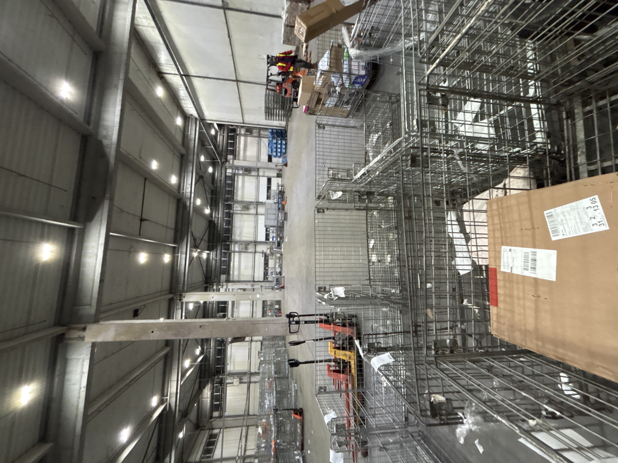 A wide-angle shot depicts the interior of a large warehouse, likely in an industrial area. The architecture consists of a high ceiling with a repetitive pattern of support beams and ceiling panels. Rows of fluorescent lights illuminate the space. The floor is smooth concrete. The foreground is dominated by stacks of wire mesh baskets. In the background, a forklift is visible, along with rows of pallet racks loaded with goods. In the midground, there are several pallet jacks. No people are clearly identifiable. The location is Burgas, Bulgaria.