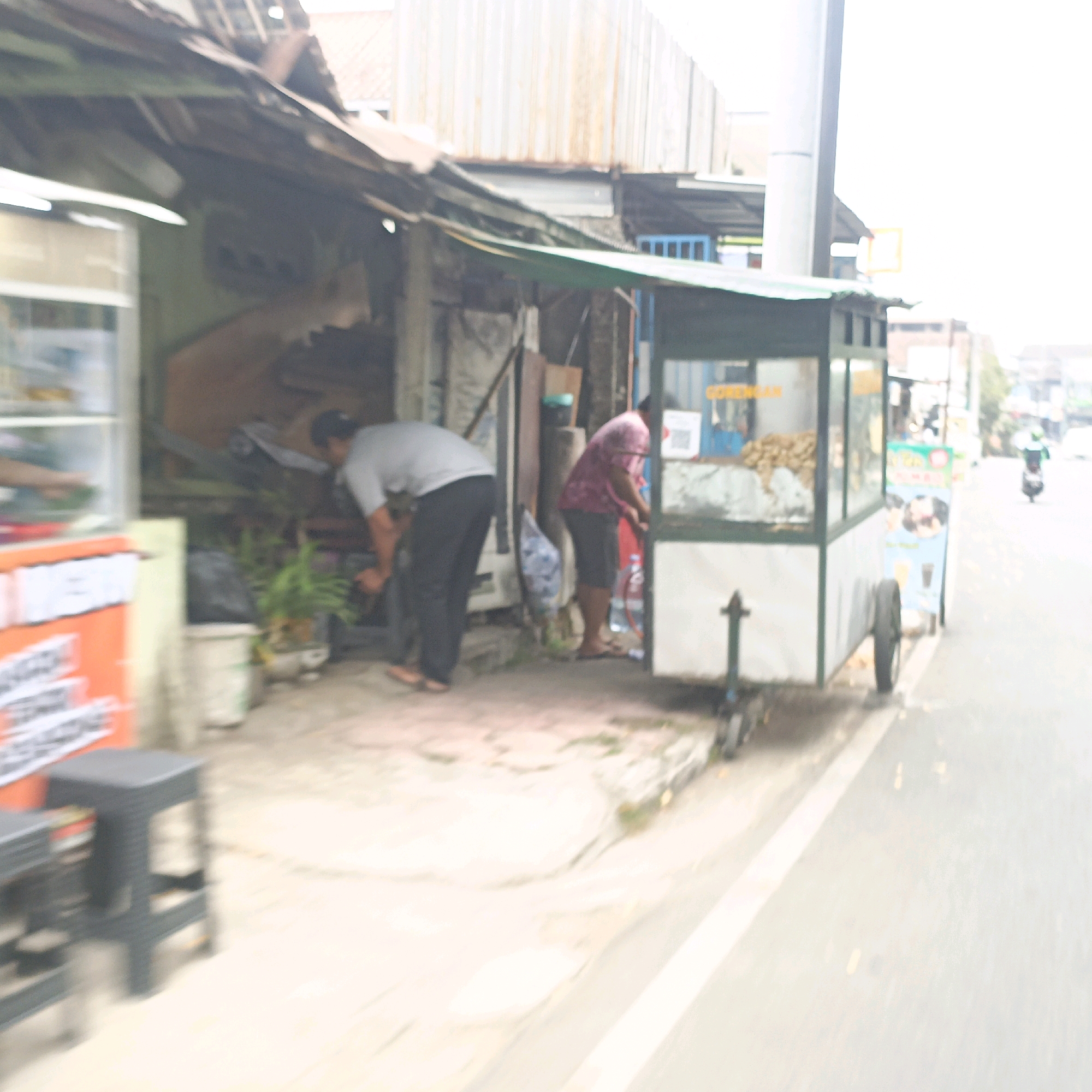 The image depicts a street scene in Salatiga, Indonesia, with a focus on a small food stall. Two individuals are visible near the stall. One person, wearing a grey shirt and dark pants, is bent over, appearing to be attending to something on the ground. The second person, in a pinkish shirt and dark shorts, is also near the stall, with their hands engaged in an activity, possibly preparing food or serving a customer.
The food stall itself is a mobile cart with a glass display case showing food items, possibly fried snacks, and a sign with yellow text, likely indicating the food being sold, such as "Gorengan". The stall is parked on a paved street or sidewalk, with a grassy area and some plants to the left of the stall. A dark-colored plastic stool is placed in front of another stall or display to the left. In the background, other buildings and a street with a motorcycle passing by are visible, suggesting an urban or semi-urban environment. The overall lighting suggests daytime.