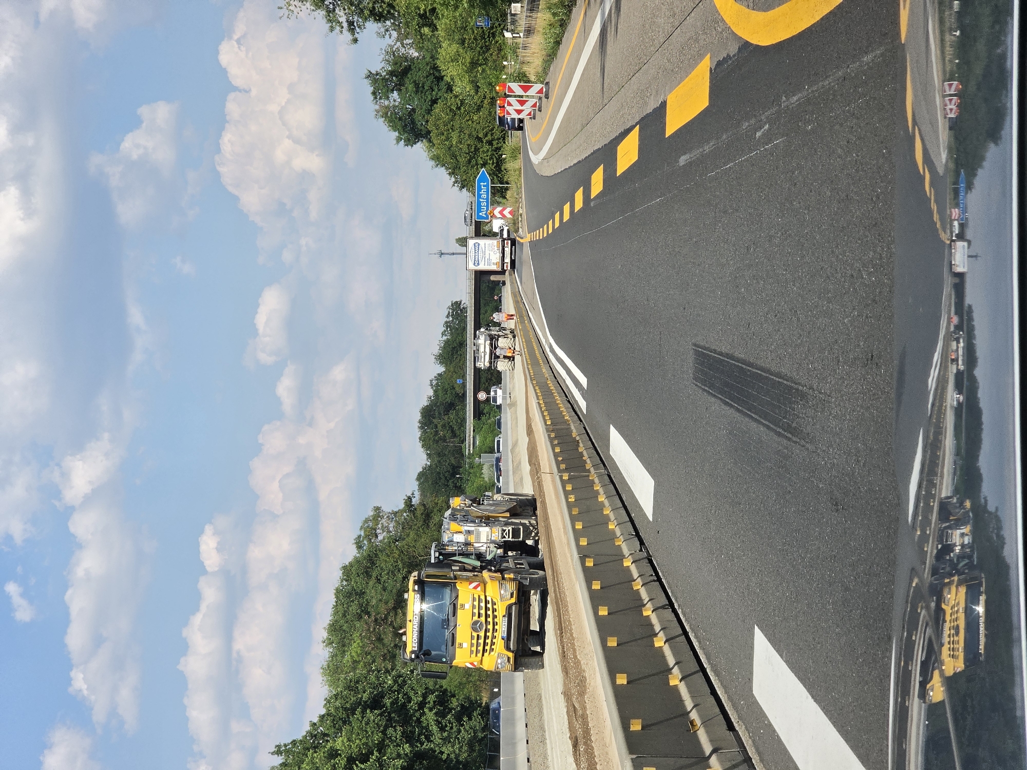 The image depicts a construction zone on a highway in Rüsselsheim am Main, Germany. A large yellow road milling machine, branded "LEONHARD WEISS," is actively working on the road surface. The machine appears to be removing material from the existing asphalt. In the background, other vehicles, including cars and trucks, are visible on the highway, some of which are navigating through the construction area. A blue directional sign indicating "Ausfahrt" (Exit) is present, suggesting an upcoming exit from the highway. The scene is characterized by temporary concrete barriers lining the construction area, road markings, and traffic cones, all typical elements of highway construction. The sky is blue with scattered white clouds, and lush green trees line the sides of the highway. The reflection on the dark surface in the foreground suggests the photo was taken from a vehicle.