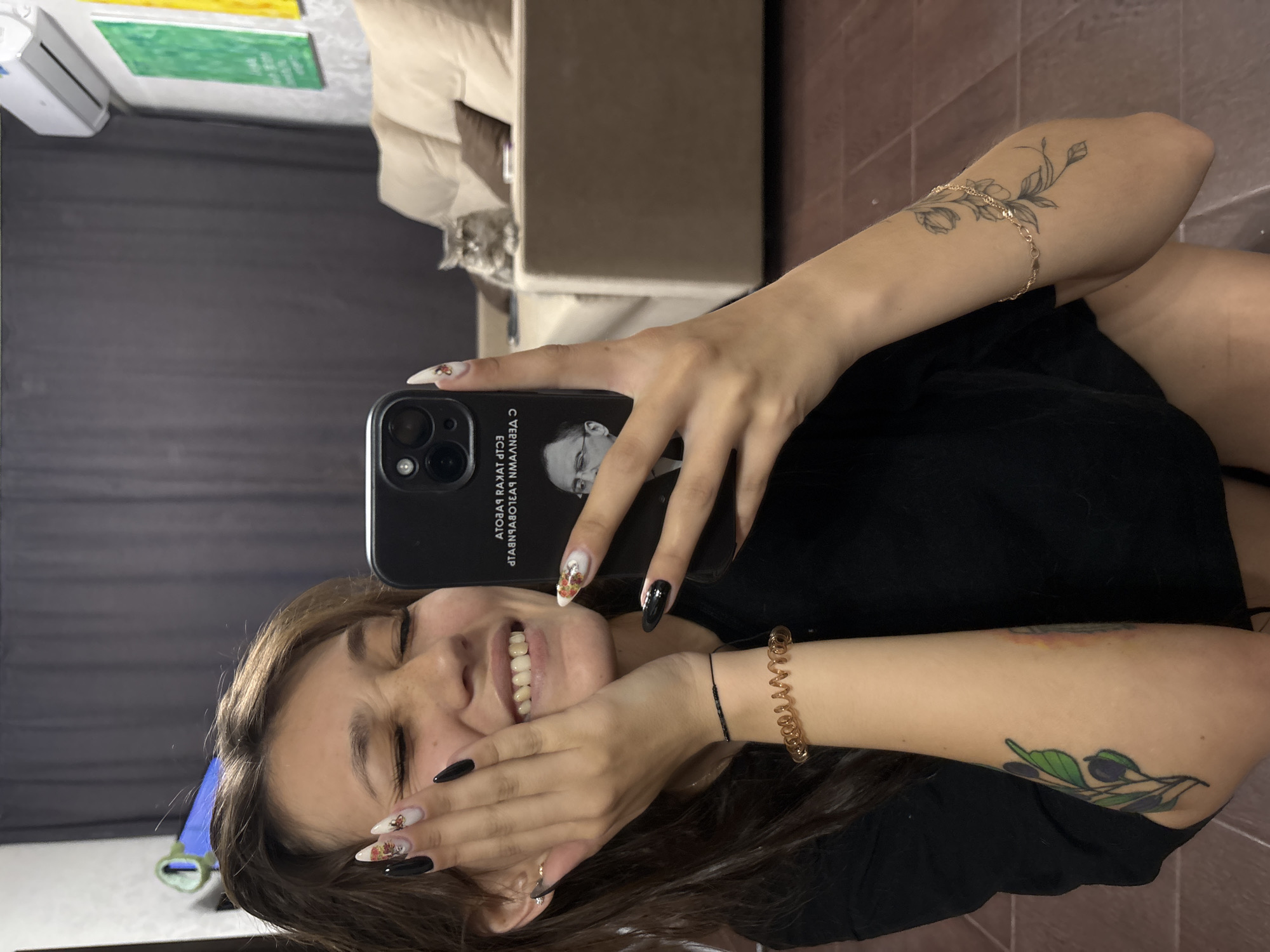 A young woman with long, dark hair takes a selfie with her smartphone. She is wearing a black t-shirt and has long, pointed fingernails with intricate designs. She is smiling widely, with her eyes closed and her left hand touching her cheek. She is wearing two bracelets on her left wrist, one a thin black cord and the other a coiled golden band. There are tattoos on her forearms; one appears to be an olive branch with black olives and green leaves, and the other is a floral design in black ink. The background shows a living room with a dark grey curtain, a beige sofa with pillows, and two framed pictures on the wall, one green and one yellow. A grey cat is visible sitting on the sofa. A white air conditioner unit is mounted high on the wall. The floor appears to be tiled or wooden. The smartphone she is holding has a black case with a picture of a man in glasses and some text, possibly in Russian. The location is not discernible from the image.