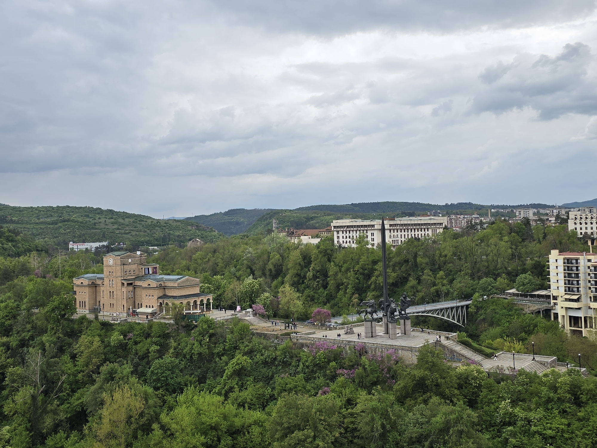 The image shows a cityscape of Veliko Tarnovo, Bulgaria, under a cloudy sky. The scene is dominated by lush green vegetation, with trees covering the slopes and hills. Several buildings are visible, including a large brick building with a clock tower, a white multi-story building, and a more modern building with balconies. A monument with statues of horsemen stands prominently on a plateau, with a bridge extending from it. People can be seen walking on the pathways around the monument and the bridge. The overall impression is one of a city built into and surrounded by a green, hilly landscape.