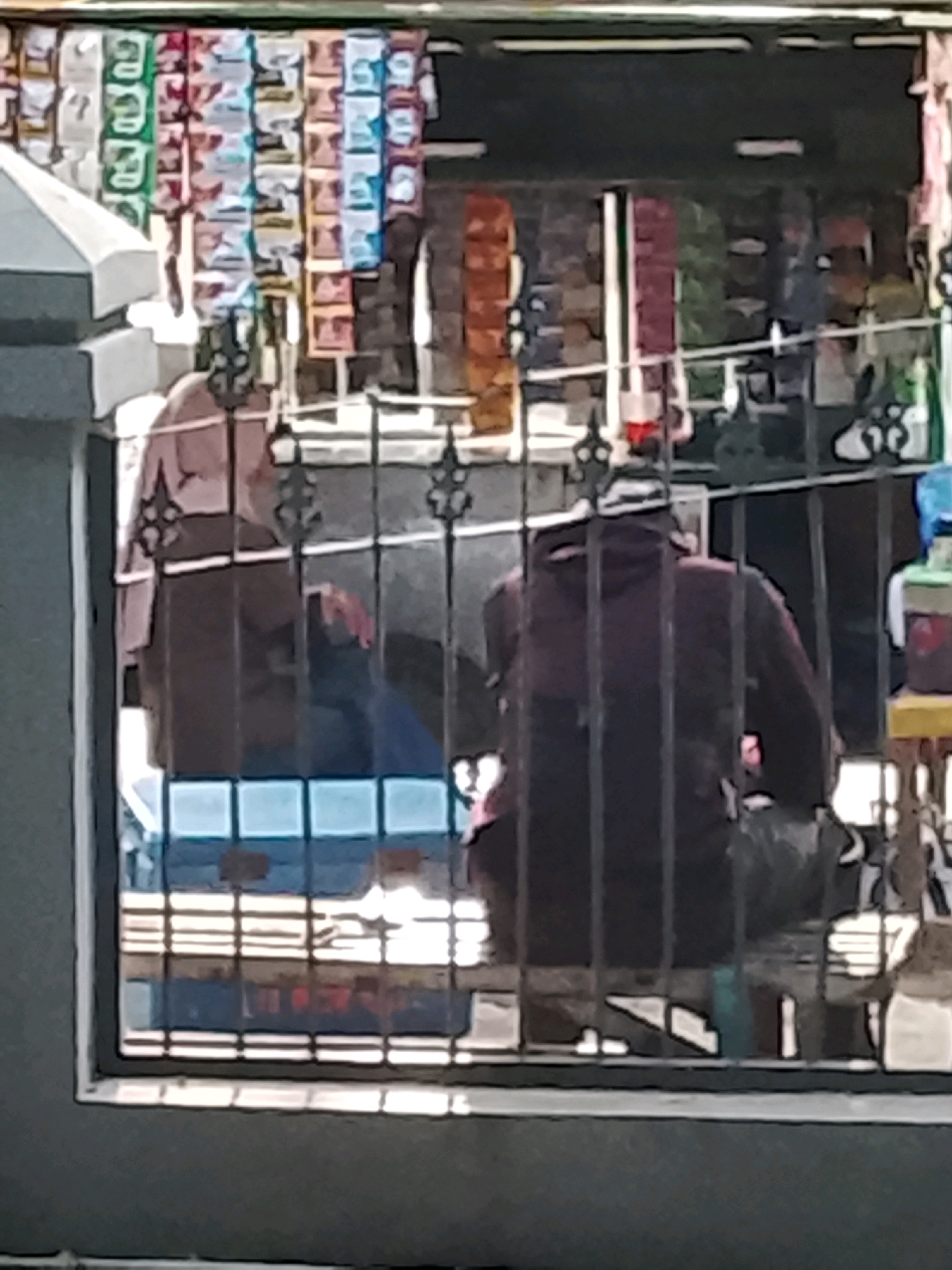 The image depicts a storefront, likely a small retail shop or "warung," in Slawi, Indonesia, viewed through a metal grate. The storefront is filled with shelves displaying numerous brightly colored items, possibly snacks or small goods, arranged in vertical rows. Two individuals are visible inside the shop, partially obscured by the grate. On the left, a person wearing a light-colored headscarf is seated. To their right, another individual is seated, wearing a dark hooded jacket. The interior of the shop appears to be dimly lit. Outside the shop, a section of a grey concrete wall is visible on the left, and a portion of a yellow stool can be seen on the right. The overall impression is of a street-level commercial establishment in a densely populated area.