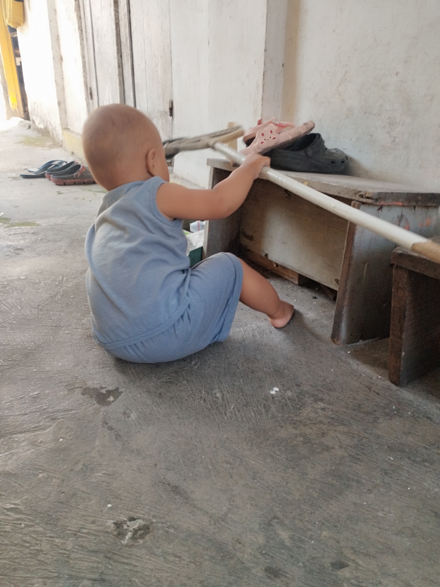 The image depicts a young child, appearing to be a toddler, sitting on a concrete floor and reaching for a broom handle that rests on a wooden structure. The child is wearing a light blue sleeveless outfit. The environment appears to be an outdoor or semi-outdoor space, possibly a pathway or yard adjacent to a building with white walls and a wooden door. Several pairs of flip-flops and clogs are visible in the background, some on the ground and others on the wooden structure. The concrete floor shows some discoloration and possibly water marks. The lighting suggests it is daytime, with sunlight casting shadows. The overall scene is simple and domestic, potentially in a residential area of Surabaya, Indonesia.