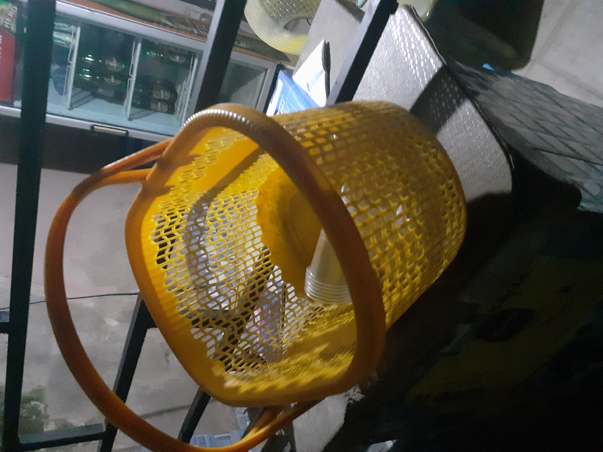 In Jalingo, Nigeria, a yellow plastic basket is placed on a dark, textured surface. The basket contains a clear plastic cup. In the background, a white refrigerator is visible with several green bottles inside. A black metal railing runs horizontally across the upper portion of the image, and a light-colored wall is visible beyond it. The scene appears to be outdoors or in an open-air setting.