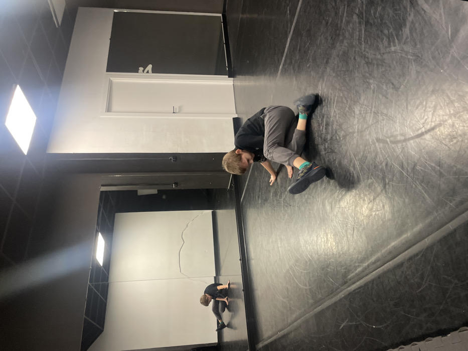 A young boy with blond hair, wearing a black shirt, gray pants, and sneakers, is on all fours on a dark, shiny floor. He seems to be in a dance studio, with a large mirror reflecting his pose. The wall has a dark section above a white section, and there are bright lights on the ceiling. The image is rotated, with the boy appearing to be crawling on a wall.