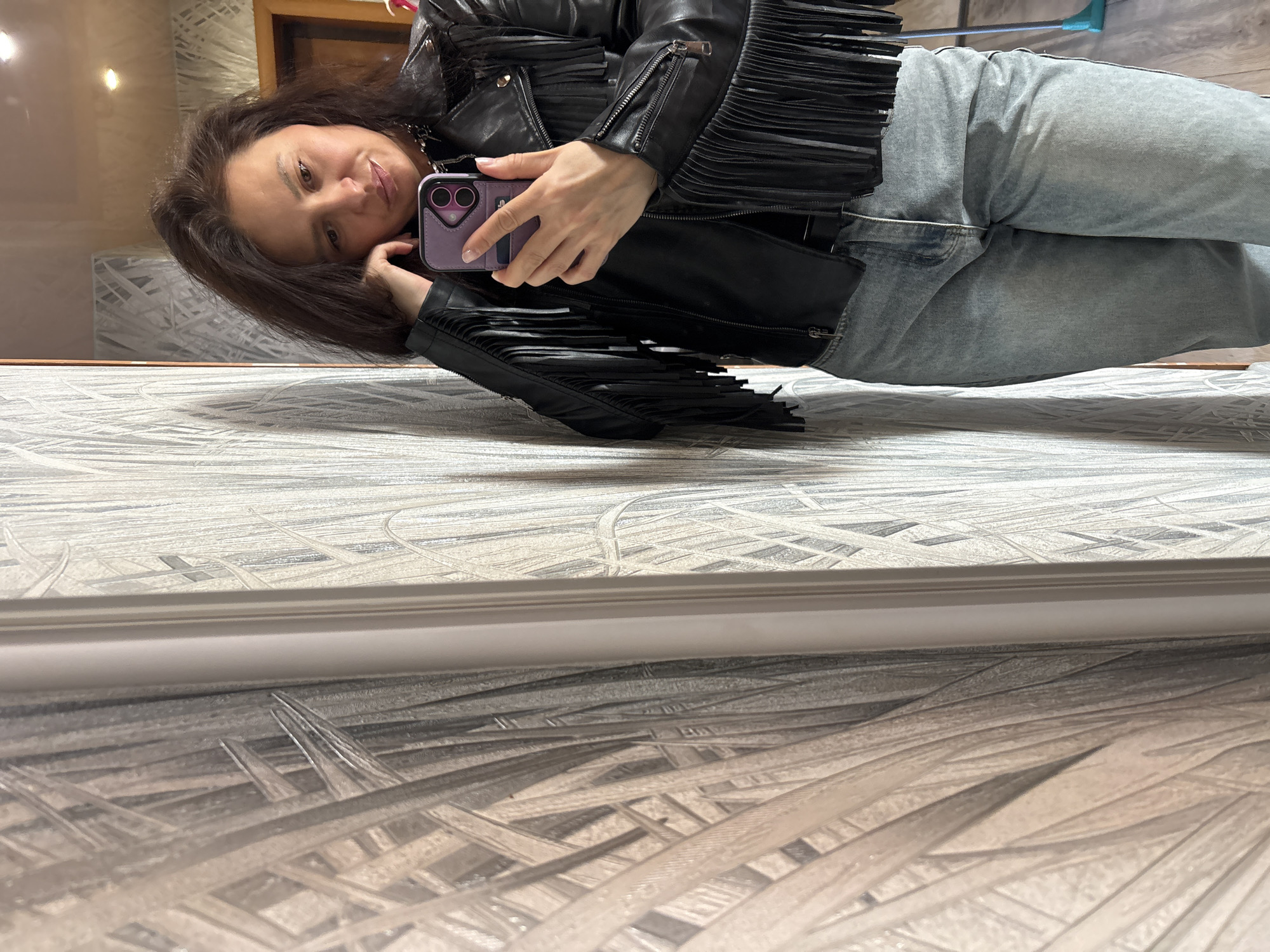 A mirror selfie captures a young to middle-aged woman reflected in a mirror, appearing to be lying or reclining. She has dark hair, a light complexion, and a gentle, pleasant smile while looking towards the camera. She holds a purple smartphone in her left hand.
The woman is wearing a black leather jacket with extensive fringe detailing on the sleeves and across the chest and back, paired with light-wash blue jeans. A silver chain necklace is visible around her neck.
The background, visible as a reflection behind her, shows elements of an indoor setting. This includes a wooden door frame, a light fixture, and walls with a light-colored, geometric or abstract patterned wallpaper. On the far right of the reflection, a blue-handled cleaning tool is faintly visible. The surface she is resting on, or the floor in front of the mirror, has a textured pattern resembling light-toned linoleum or a rug with abstract, interwoven lines.
The activity is the woman taking a self-portrait indoors, likely in a private setting such as a home in Chaikovsky, Russia. Her expression suggests a moment of contentment. No discernible text is visible in the image.