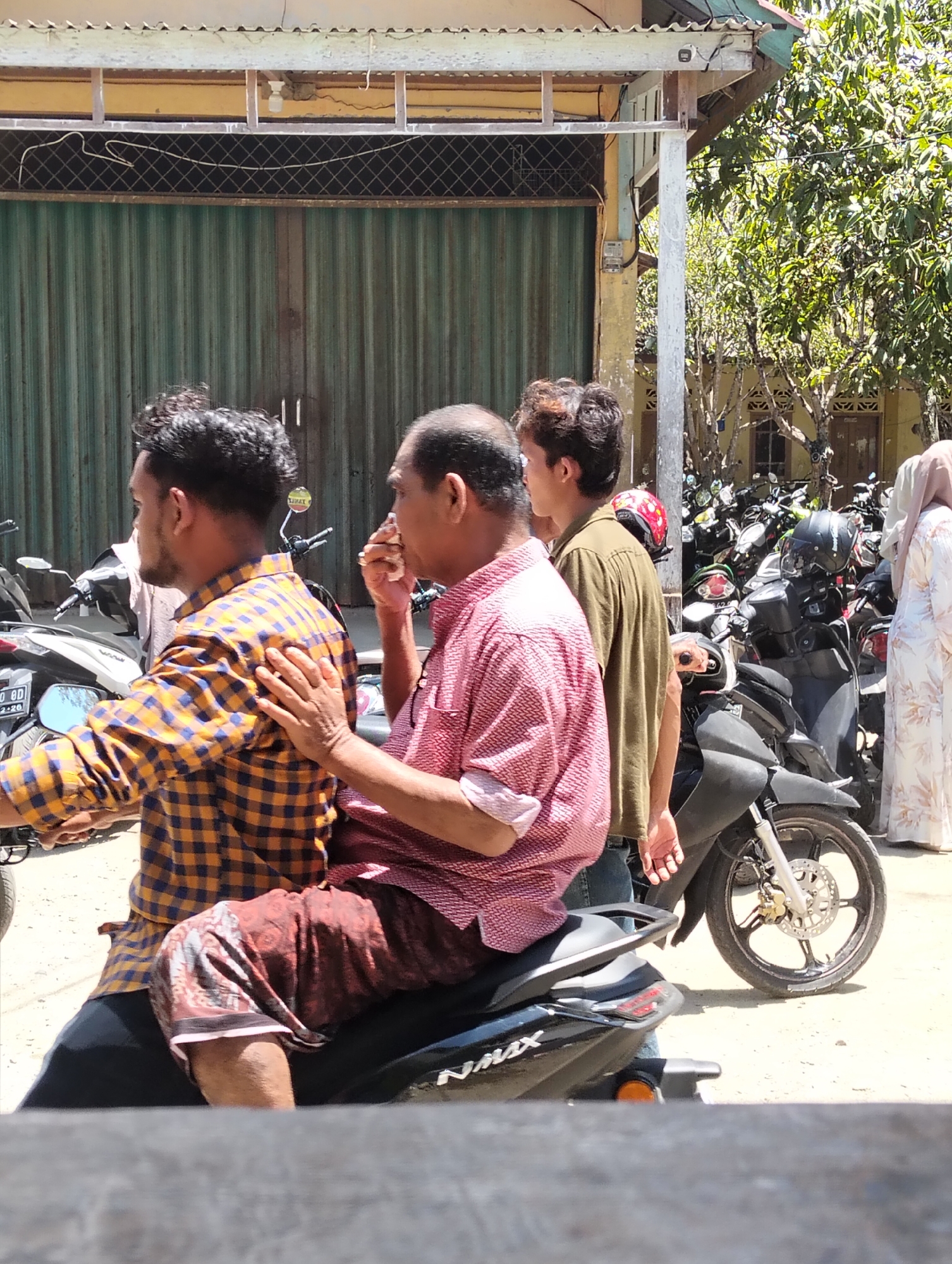 In Teungoh, Indonesia, a bright, sunny day illuminates a scene with three men and one woman. Two men are seated on a dark-colored motorcycle in the foreground. The driver, wearing a yellow and blue checkered shirt, has his back to the camera, while the passenger, in a pink checkered shirt, sits behind him and appears to be wiping his nose with his right hand. Another man, wearing a green shirt, stands to the right of the motorcycle. A woman, dressed in a light-colored patterned dress and hijab, stands further back, to the right of the group. In the background, a large gathering of parked motorcycles is visible, along with a building featuring a corrugated green metal door. The ground is covered in a light-colored, dusty surface.