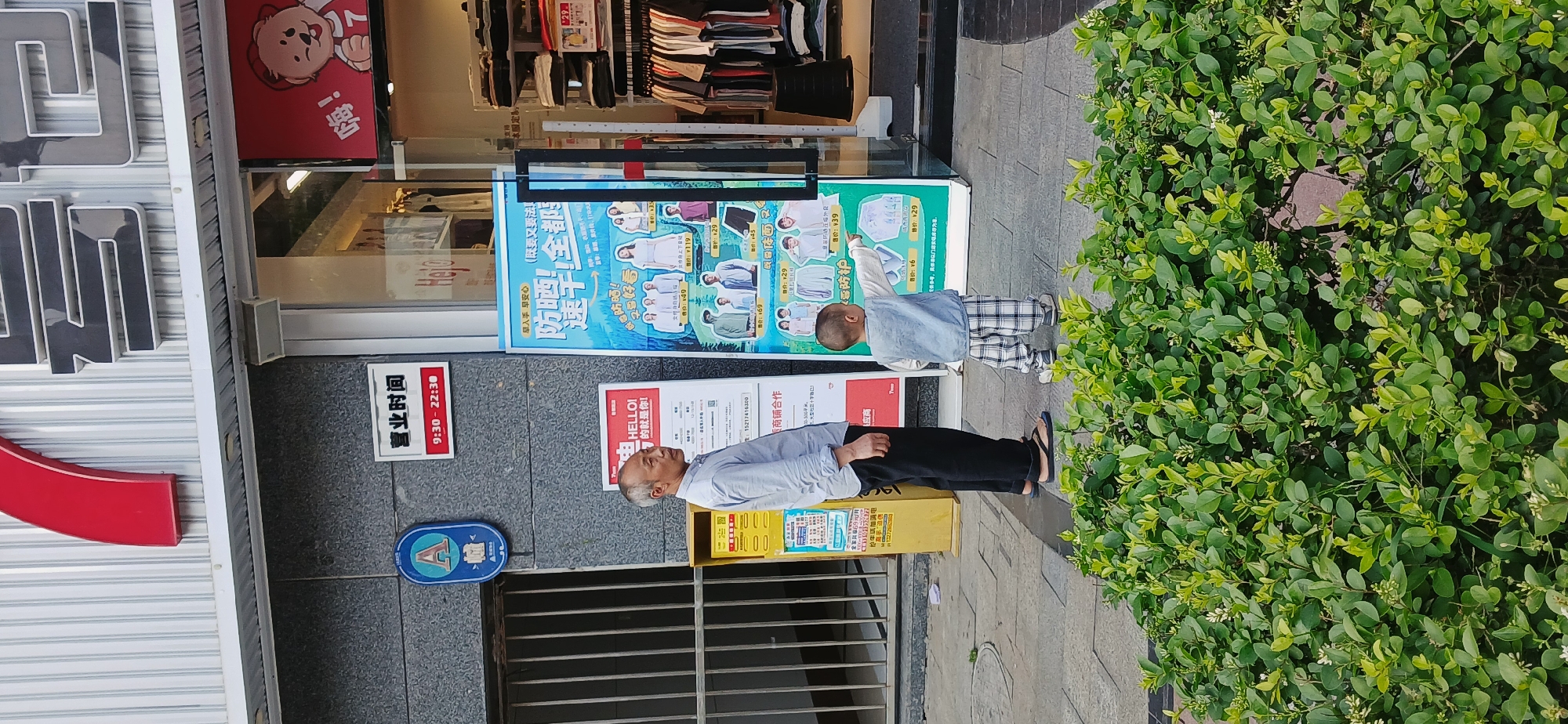 Here's a detailed description of the image:
The photo captures a street scene, possibly in Shapingba District, China, based on the visible Chinese signage. An elderly man in a light-colored shirt and dark pants stands near a store, appearing to look at a poster or advertisement display. Beside him, a young child wearing a denim vest over a light-colored top and plaid pants points towards the store's display. The display features images of clothing items and pricing in Chinese Yuan.
The store itself has a glass front where clothing is visible inside. The store front has red signage with a cartoon bear on it, as well as signage indicating the store's opening hours from 9:30 to 22:30. A sign with the phone number 15217416300 is present.
Next to the shop entrance are some green bushes and a drain cover. The ground is paved with grey stone.
