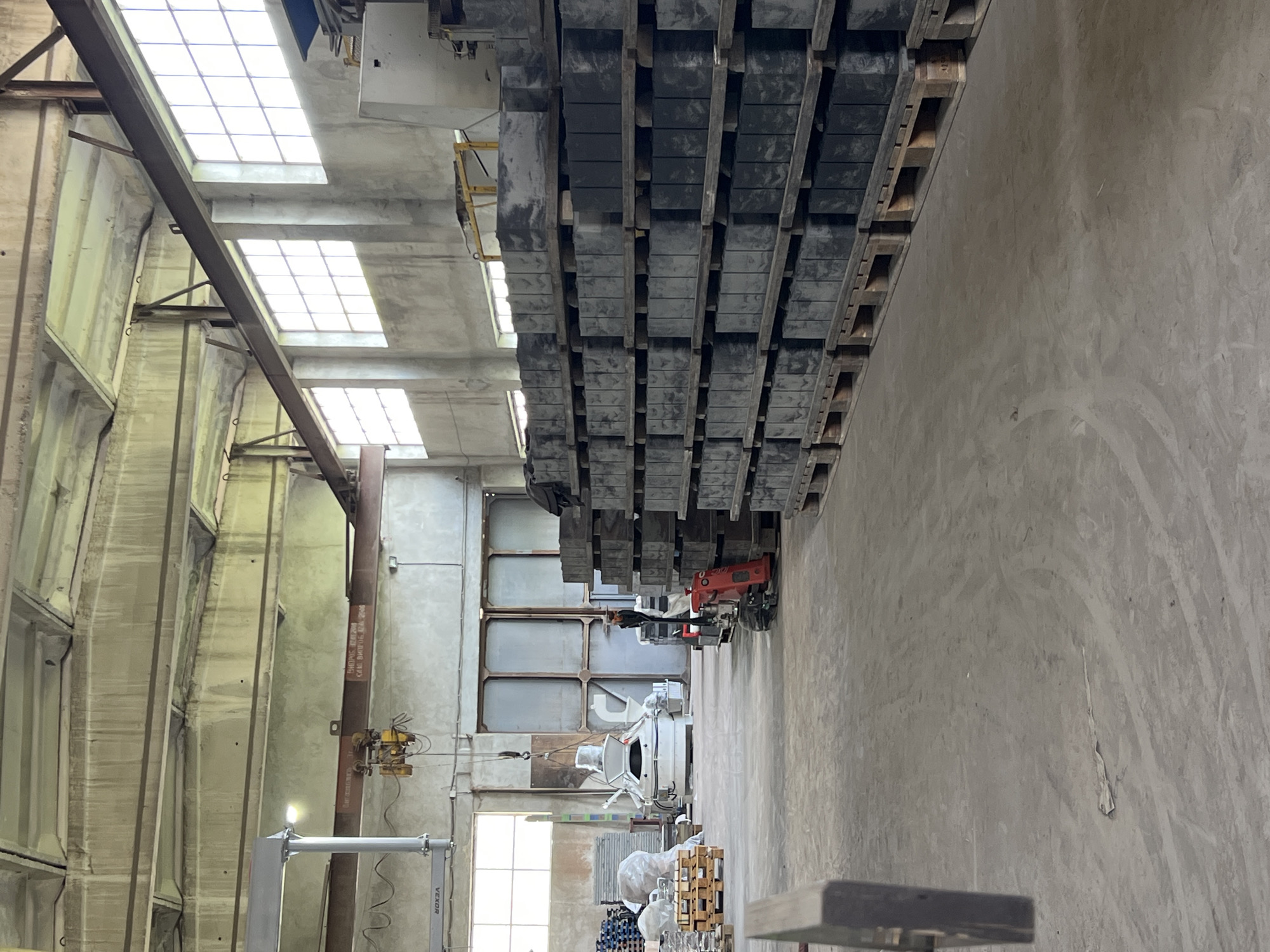 The image depicts the interior of what appears to be a large industrial building, possibly located in Uhry, Ukraine, though this is not directly verifiable from the image. The building features high ceilings with multiple skylights, steel support beams, and concrete walls and floors. Stacks of dark gray rectangular objects, potentially concrete blocks or pavers, are palletized and stacked high, dominating the central foreground. A forklift is visible among the stacked pallets, suggesting material handling operations. Further into the background, there are other industrial equipment and materials, including stacks of wooden pallets. There are no people visible in the image. The scene suggests a manufacturing or storage facility.