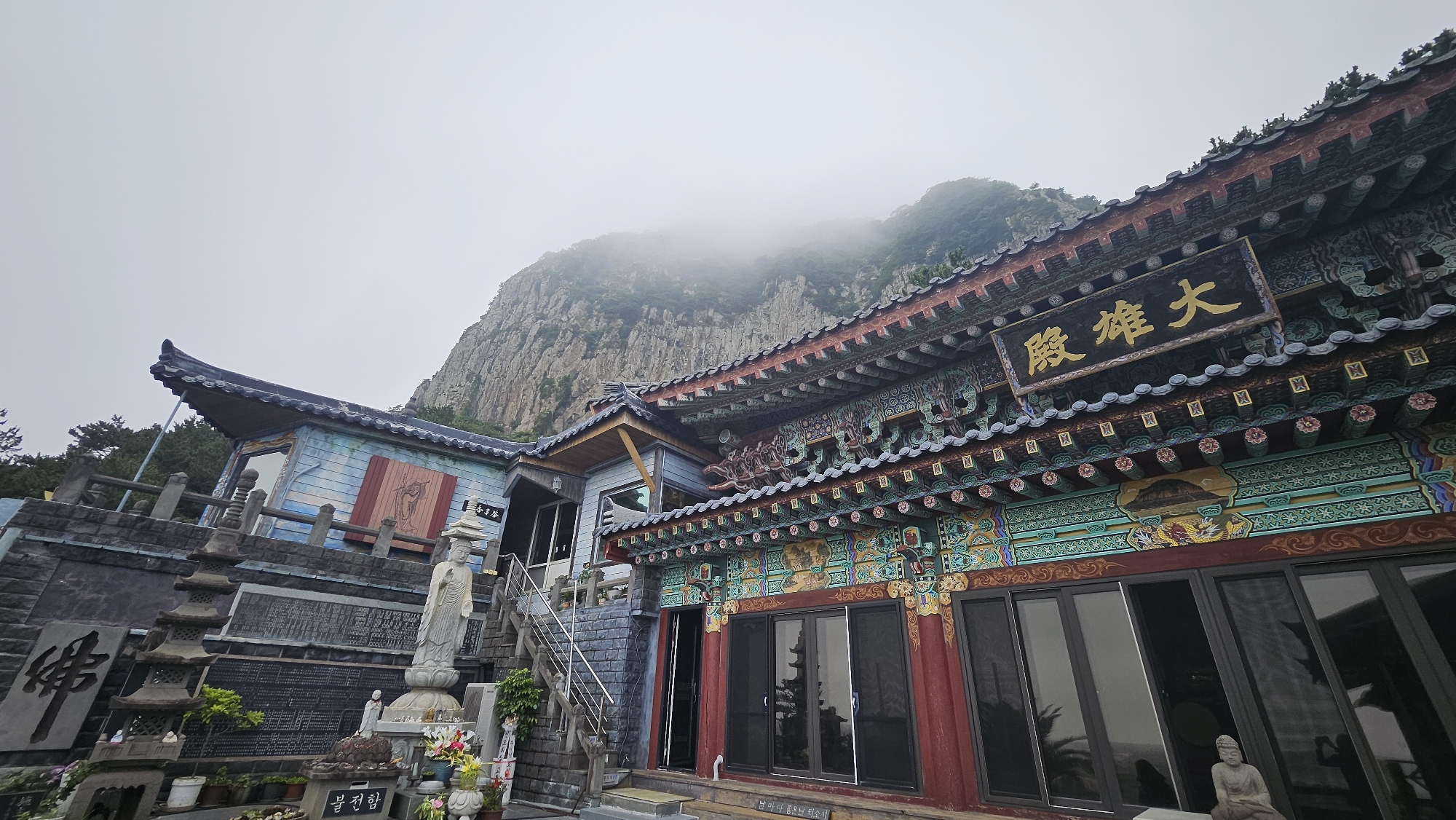 The image captures a section of a temple complex situated in Seogwipo-si, South Korea. The temple features traditional East Asian architectural elements with ornate, colorful decorations on the eaves and facades. A large sign with Korean characters in gold is prominently displayed on one of the buildings. In the foreground, there are stone stupas and statues, including a standing Buddha figure. A steep, rocky mountain shrouded in mist looms in the background, indicating a mountainous or cliffside location within the city. The sky is overcast, contributing to a somewhat muted atmosphere.