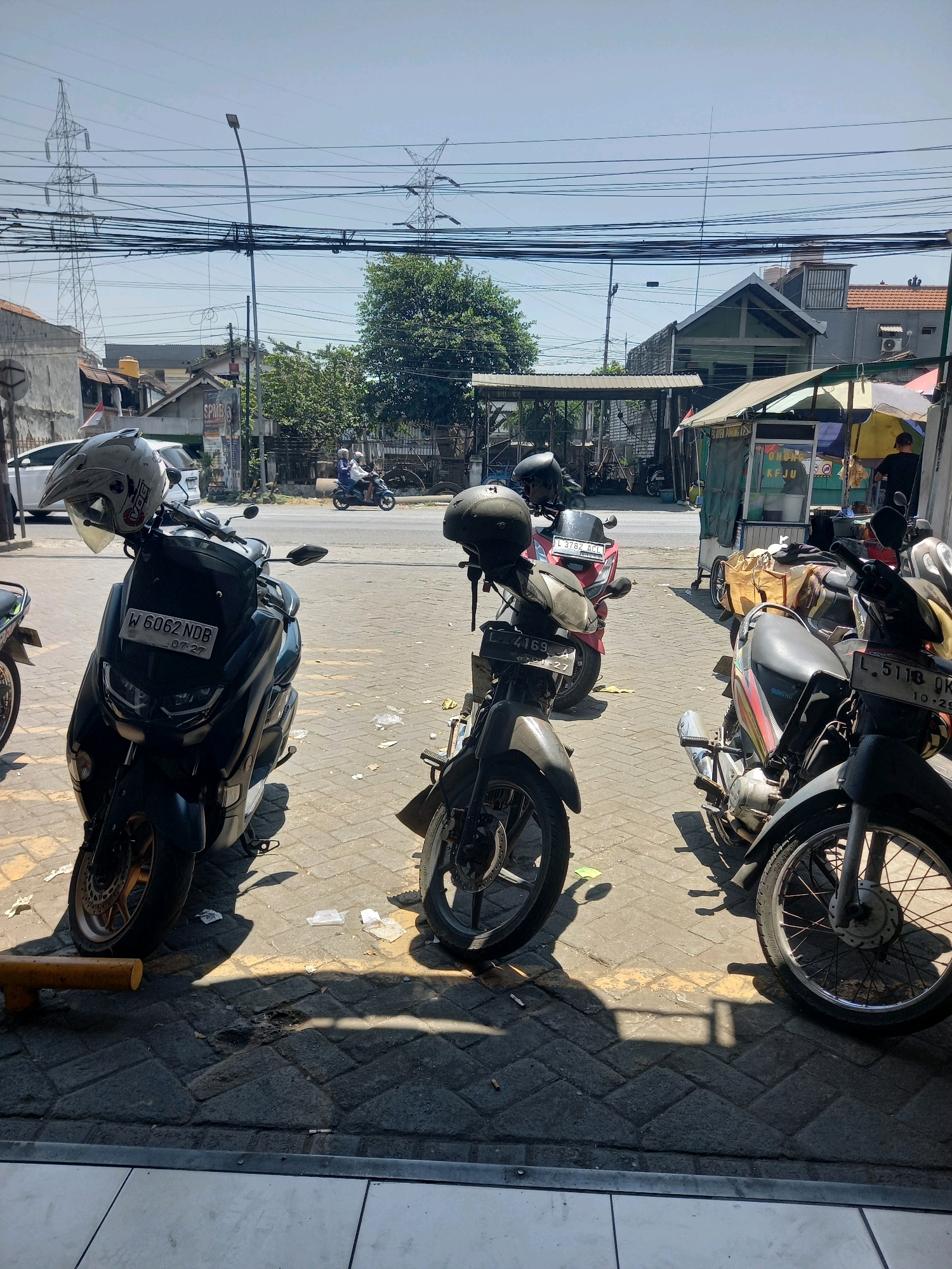 In Surabaya, Indonesia, this outdoor scene captures a street-level view in front of a commercial establishment, possibly a food stall or shop. Several motorcycles are parked on a paved ground of interlocking concrete bricks. The foreground features a dark blue scooter with a white helmet resting on its handlebars. To its right, a silver and grey motorcycle displays a license plate that reads "L 5118 OK" with a date below. Another dark-colored motorcycle to the left of the blue scooter is partially visible. In the mid-ground, a red scooter with license plate "L 3782 ACL" is parked, followed by another motorcycle and further down, a red and white scooter. Behind the motorcycles, a low-lying structure with a green awning extends across the scene. A person in a dark shirt can be seen standing behind a counter in the stall. In the background, power lines crisscross the sky, supported by tall transmission towers. The sky is clear and blue, indicating a sunny day. The street beyond the parked vehicles shows a few more motorcycles, and traffic can be observed in the distance. The overall impression is a busy urban environment with a focus on personal transportation.