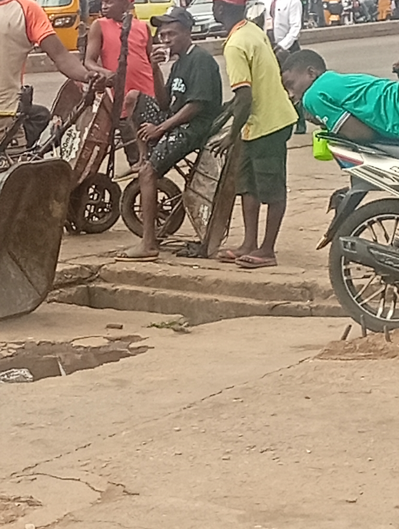 The image depicts a street scene in Maiburiji, Nigeria. In the foreground, there is a cracked concrete surface with a large, open hole. Several individuals are positioned on a slightly elevated platform in the background. A man wearing a black shirt and cap sits on a modified, tricycle-like vehicle, holding a cup to his mouth. Another man, wearing a yellow shirt, stands near the tricycle. A third man in a green shirt is lying across the seat of a parked motorcycle. Additional individuals and vehicles are visible further in the background, including a yellow auto-rickshaw. The overall scene suggests a bustling urban environment with visible infrastructure issues.