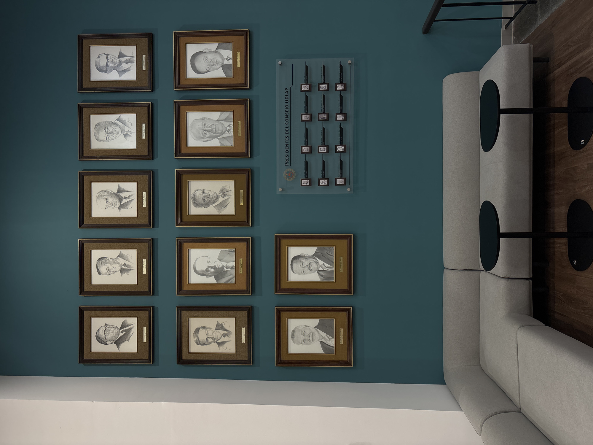 The image depicts a wall in a room, potentially located in the city of San Andrés Cholula, Mexico. The wall is painted in a teal-blue color and adorned with several framed portraits arranged in rows. The portraits appear to be sketches or drawings of men. Adjacent to the portraits, there is a clear plaque with the inscription "Presidentes del Consejo UDLAP". The plaque includes additional smaller portraits and associated text. To the right of the wall, there is a grey L-shaped padded seating arrangement with small, round black tables. The floor is made of wood. The overall setting suggests an institutional or memorial space, possibly within the Universidad de las Américas Puebla (UDLAP), given the mention of "Consejo UDLAP". The location of the city is inferred from the presumed location of the university. The room appears to be tidy and well-maintained.