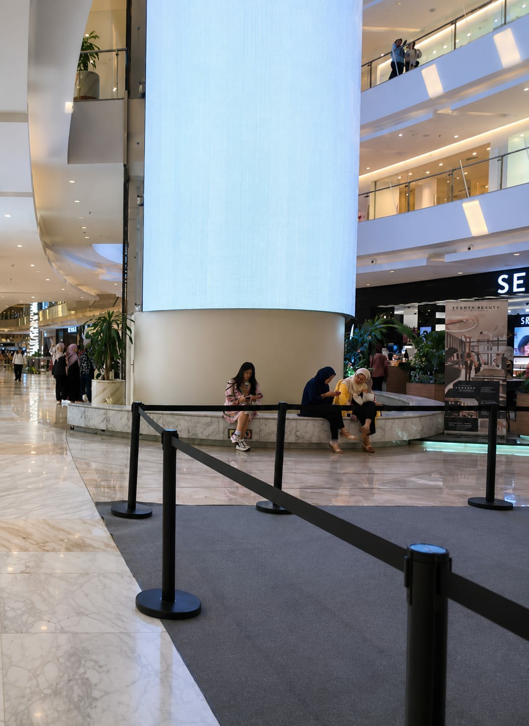 The image depicts a wide shot of a brightly lit, multi-level indoor shopping mall in the city of Medan, Indonesia. The central focus is a massive, cylindrical, light-up display column that dominates the foreground and mid-ground. In front of this column, a marble-lined bench is occupied by three young women. The woman on the left is wearing a pink and white patterned dress and is looking down at her phone. The woman in the middle is wearing a blue hijab and dark clothing and is also looking at her phone. The woman on the right is wearing a yellow hijab and white clothing and is conversing with the woman next to her. A black velvet rope barrier with stanchions runs across the lower part of the image, creating a defined area in front of the bench.
The mall environment is characterized by polished marble floors, multiple levels connected by escalators and walkways visible in the background, and large storefronts. One prominent storefront in the mid-ground reads "SEPHORA" and displays "FENTY BEAUTY" merchandise. Lush green plants are strategically placed throughout the mall, adding a touch of nature to the man-made structure. In the background, various shoppers can be seen walking through the mall, further indicating a busy retail environment. The overall impression is one of a modern, spacious, and upscale shopping destination.