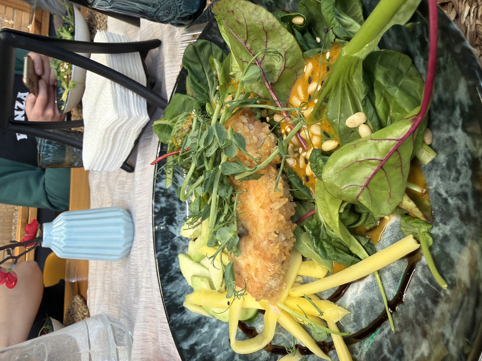 The image depicts a plate of food, possibly a salad, featuring greens, what appears to be fried fish, mango slices, and various other ingredients, placed on a dark, marble-patterned plate. There's also a light blue vase with red flowers nearby, along with napkins and a chair. A person is visible in the background using a phone to capture a shot of the food, seated at the table. The scene appears to be inside a restaurant. There is no indication of the location being in Izhevsk, Russia within the image.