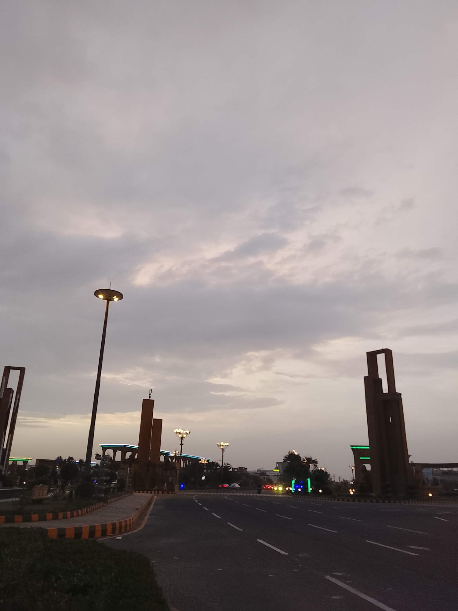 This image captures a modern urban landscape at dusk or on a heavily overcast day, set in Al Falah Modern City, Pakistan. The scene is dominated by a cloudy, pale grey sky, suggesting low natural light.
The foreground features a multi-lane asphalt road with clear white lane markings, leading into the distance. A median strip, bordered by a distinctive black and yellow striped curb, separates the traffic directions. This median is adorned with several tall, dark, rectangular architectural structures, possibly monuments or gateway elements, which give the area a contemporary aesthetic. Some of these structures have open, rectangular sections at their tops.
Artificial lighting is prominent, with a tall, modern streetlight featuring a circular, multi-lamp fixture illuminating the left side of the frame. Further down the road, other streetlights with classic globe-shaped lamps are visible. Green and blue accent lights highlight some of the distant structures and trees, adding a vibrant, modern touch to the urban design. Low-lying green bushes and shrubs line the median and the immediate foreground.
The road appears largely empty of vehicles and there are no discernible people, contributing to a tranquil atmosphere. The combination of modern architectural elements and artificial lighting against the dim, cloudy sky characterizes a well-developed urban area at twilight. No discernible text is visible within the image.