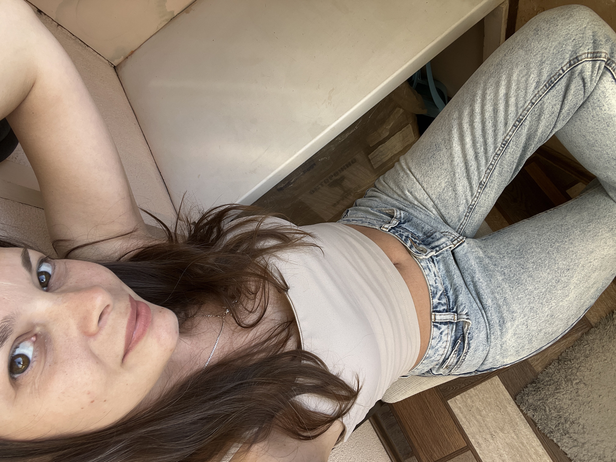 The image shows a woman lying on her side, with her head close to the camera. She has long, dark brown hair and is wearing a light-colored tank top and light-wash jeans. Her eyes are open and looking towards the camera. The setting appears to be indoors, with a light-colored surface behind her and a wooden floor visible. A rug is also visible in the lower right corner of the image. The lighting suggests it is daytime. There is no discernible text in the image.