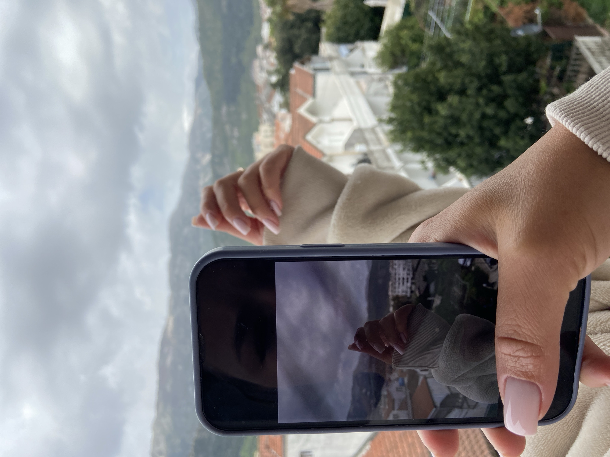 A person is holding a blue smartphone, taking a picture of their hand with the backdrop of Budva, Montenegro in the background. The person is wearing a beige colored sweater. The smartphone is partially obscuring the view. The background shows a cloudy sky and a cityscape with buildings and mountains in the distance.
