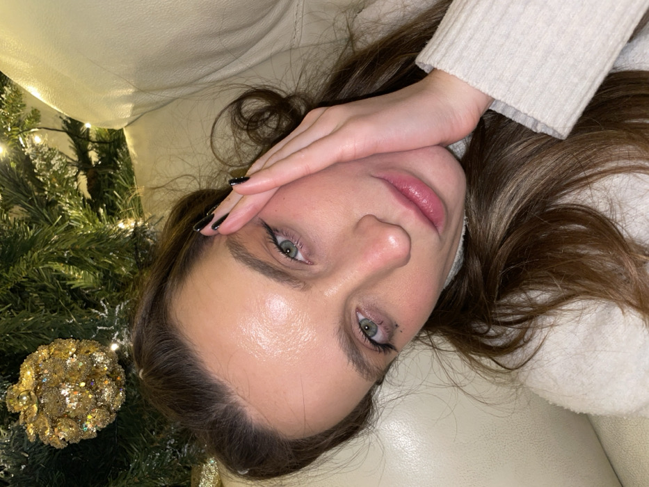 A woman with fair skin and brown hair is lying down, covering part of her face with her hand. She has light green eyes and appears to be wearing eyeliner and lip gloss. Her nails are painted black. A Christmas tree with golden ornaments is visible in the background. The woman is wearing a light-colored sweater and is lying on a beige-colored surface.