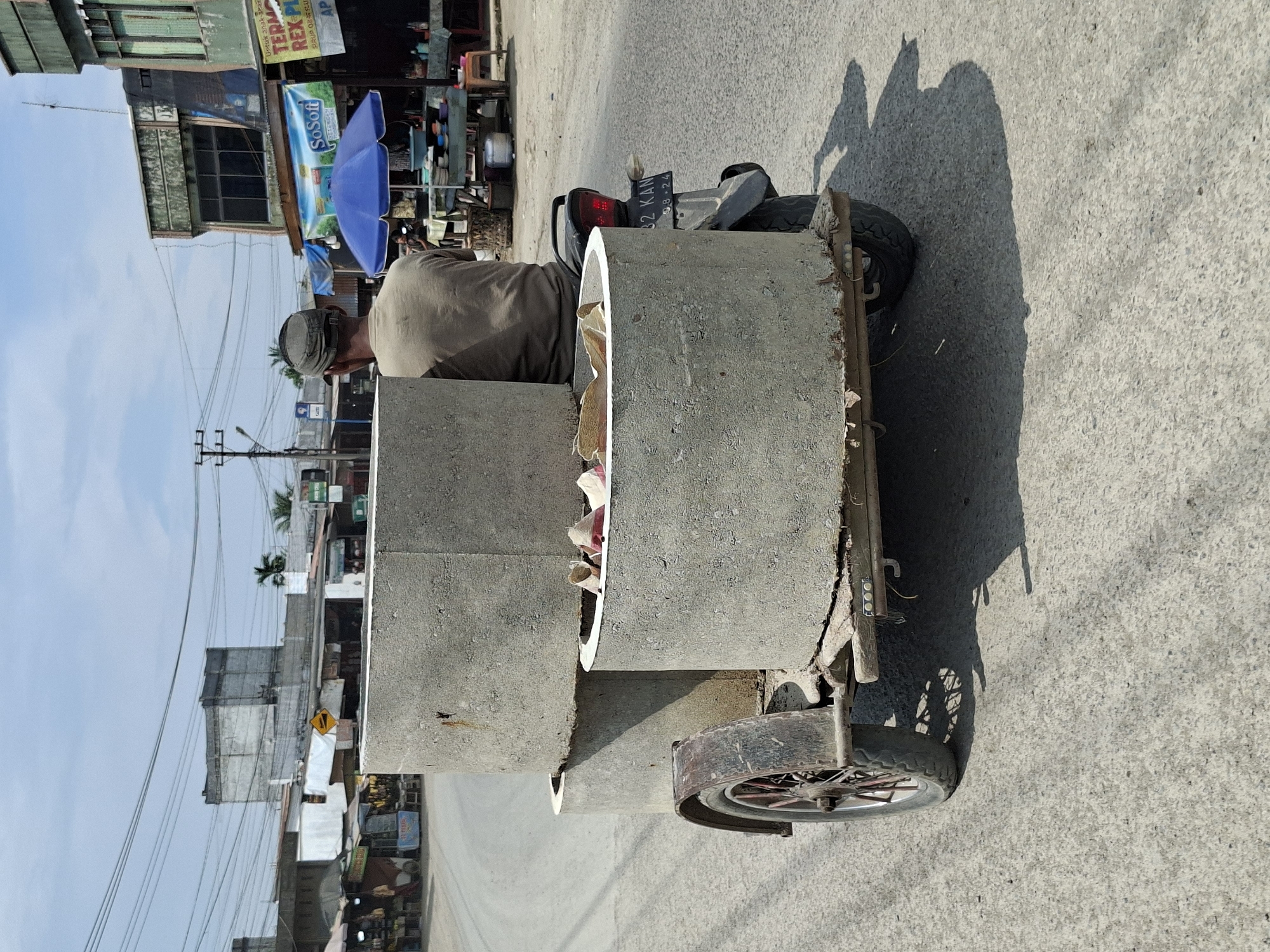 In Keudegeudong, Indonesia, a person wearing a beige shirt and a cap is riding a motorcycle that is pulling a trailer loaded with large concrete cylinders. The trailer appears to be homemade, constructed from metal and wood, with a single wheel visible on the left side. The concrete cylinders are stacked, with two full cylinders and one partially filled with what appear to be paper or fabric scraps. The motorcycle has a license plate that reads "92 KAN 08-24". The scene takes place on an asphalt road, with buildings and market stalls lining the street on the right side. Overhead wires crisscross the sky, and the weather is clear with a bright sun casting shadows. The overall impression is one of everyday commerce and transportation in a bustling Indonesian town.