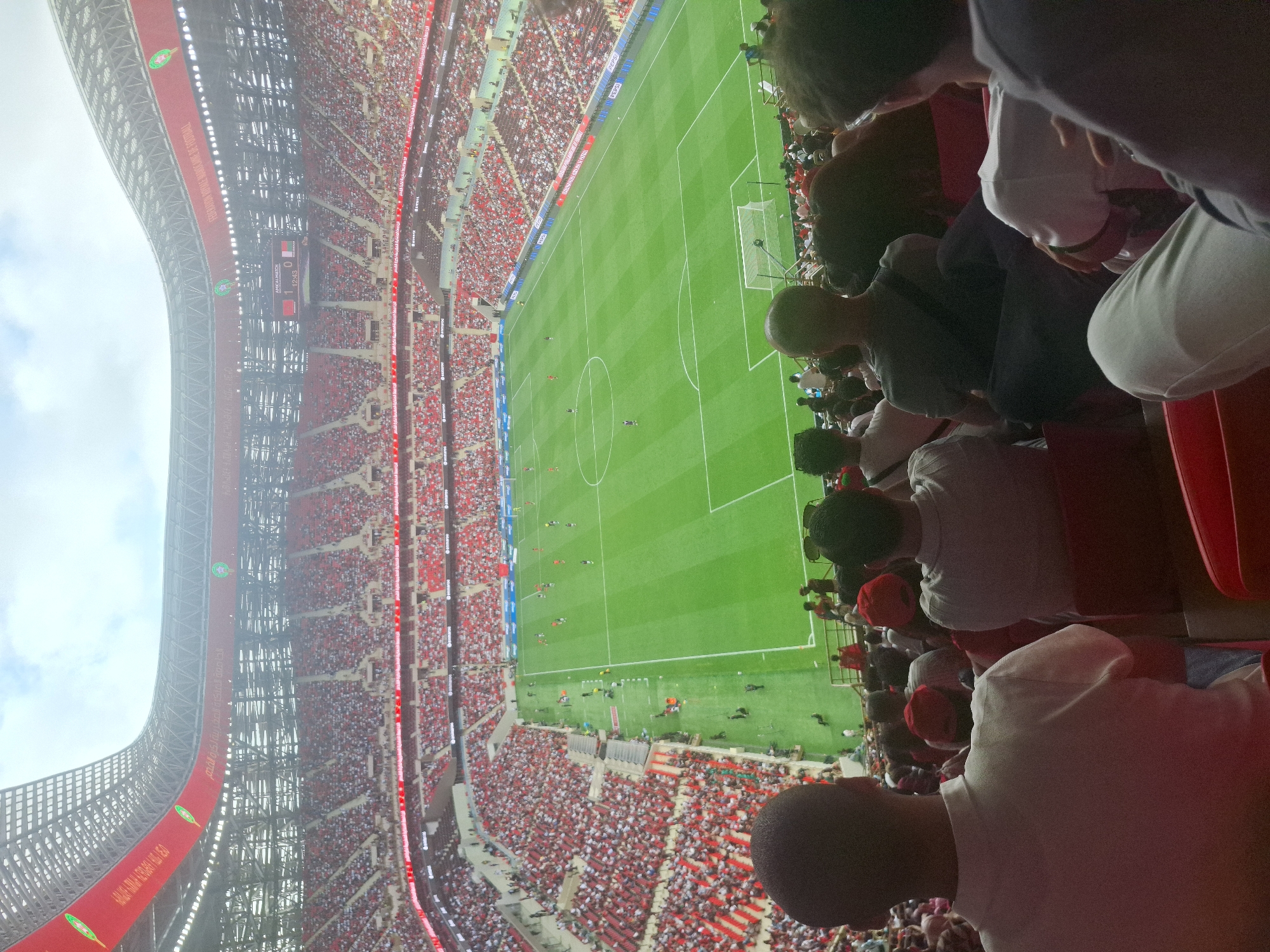 This media file captures a high-angle view of a vibrant football match in a large, modern stadium, likely located in Rabat, Morocco, as indicated by the user. The stands are densely packed with spectators, many wearing red and white, suggesting support for the Moroccan national team. Several individuals are visible in the immediate foreground, their backs to the camera, observing the game.
The green football pitch is clearly visible, with players scattered across it, indicating an active match. A scoreboard on the upper left side of the stadium displays "AMICAL MAROC" (Friendly Morocco), with a score of "1 2:43 0". The flag next to the "1" is red with a green star, representing Morocco, while the flag next to the "0" is white, green, and red vertical stripes (possibly Italy or Algeria, given the context of a friendly).
Along the stadium's structure, large banners are prominently displayed. One banner clearly reads "FEDERATION ROYALE MAROCAINE DE FOOTBALL" in French, with additional Arabic text below it. Another banner further along features the Moroccan flag emblem and more Arabic text, reinforcing the Moroccan context. Advertisements, including the Pepsi logo, are visible on hoardings around the pitch. The time of day appears to be daytime, with a clear sky visible above the stadium roof, although there's some glare or haze. The general atmosphere appears energetic, consistent with a live football event.