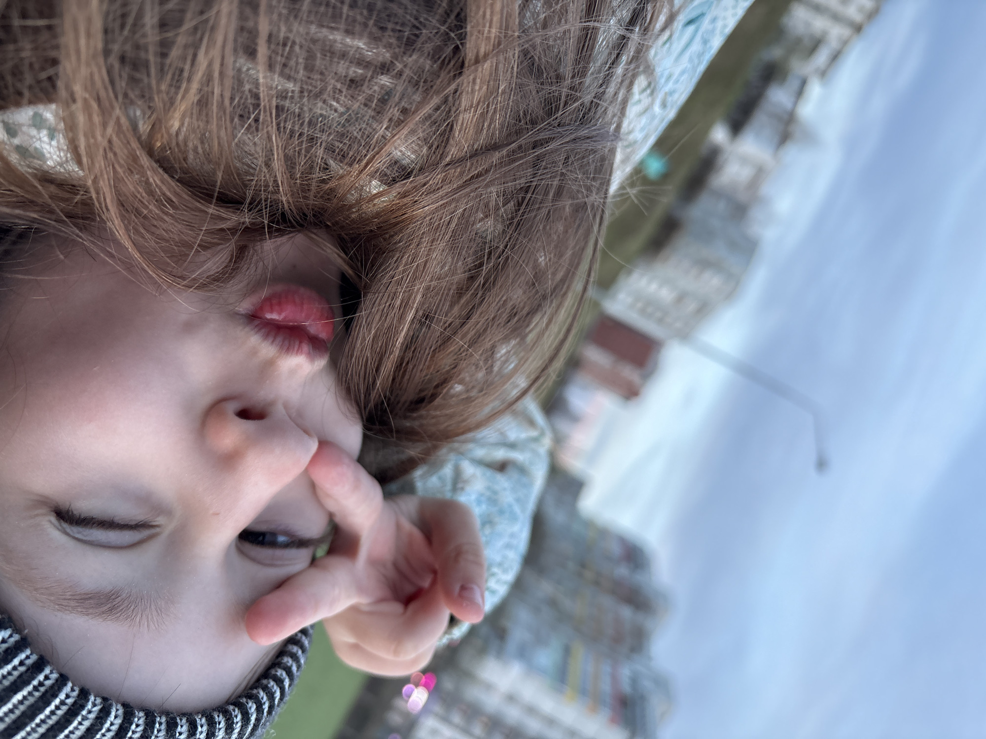 A young person, possibly a child, is depicted upside down. They are wearing a black and white striped beanie and a patterned top. Their hand is raised in front of their face, partially obscuring one eye. The background reveals a blurred urban landscape, with buildings and a sky visible. The location is identifiable as Chaikovsky, Russia, due to distinctive buildings and architecture visible in the background.