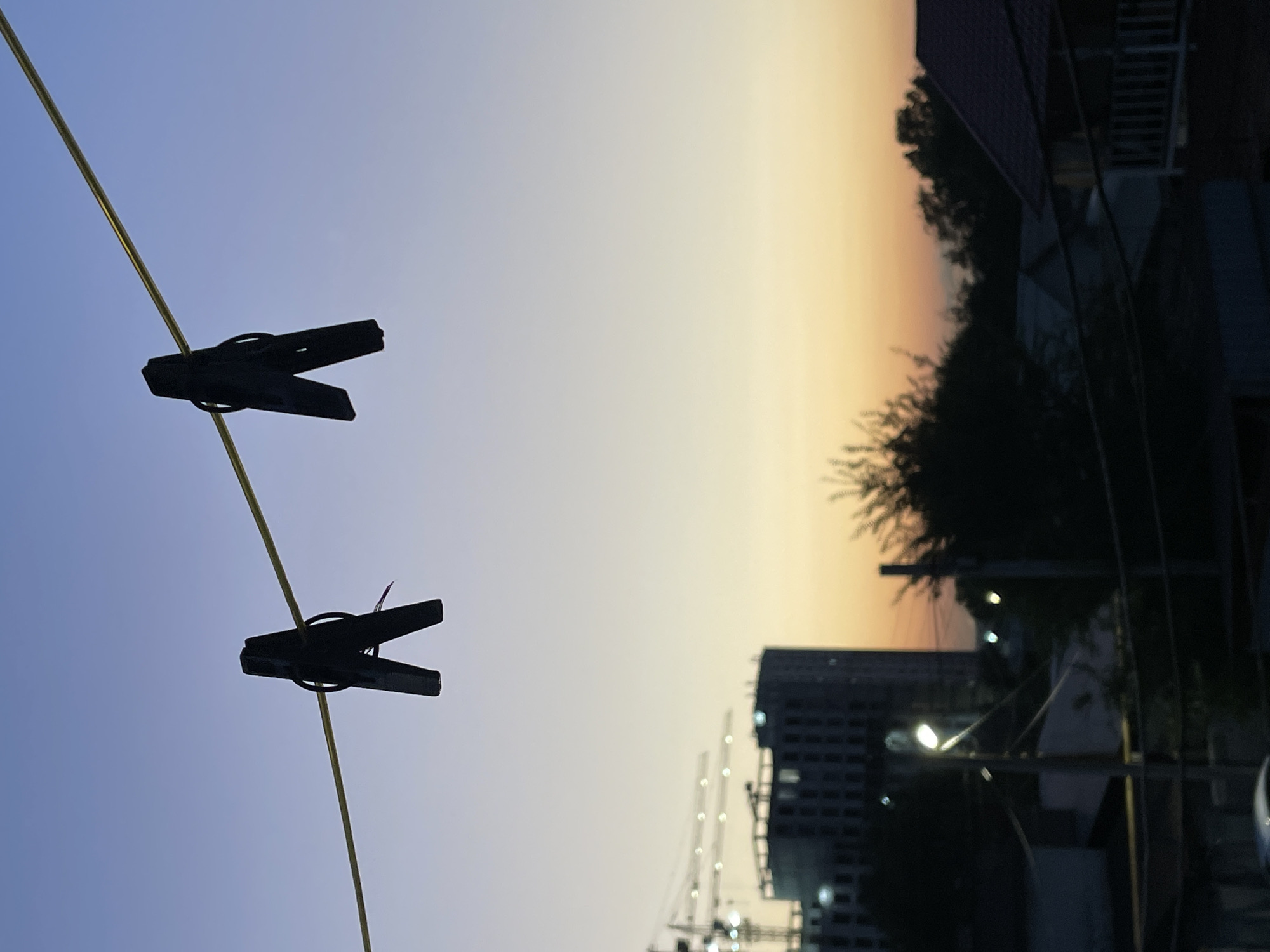 The image depicts a dusky sky with a gradient from light blue at the top to orange and yellow near the horizon. Two black clothespins are clipped to a thin, yellow clothesline that bisects the upper portion of the frame. Below the clothesline, the silhouettes of buildings and trees are visible. In the mid-ground, a construction site with cranes and a large, dark building suggests urban development. A streetlamp is illuminated, casting a soft glow. The lower portion of the image shows the rooftops of houses. The scene appears to be an outdoor view from a residential area, possibly a balcony or window, looking towards a developing part of the city. The location is identified as Almaty, Kazakhstan.
