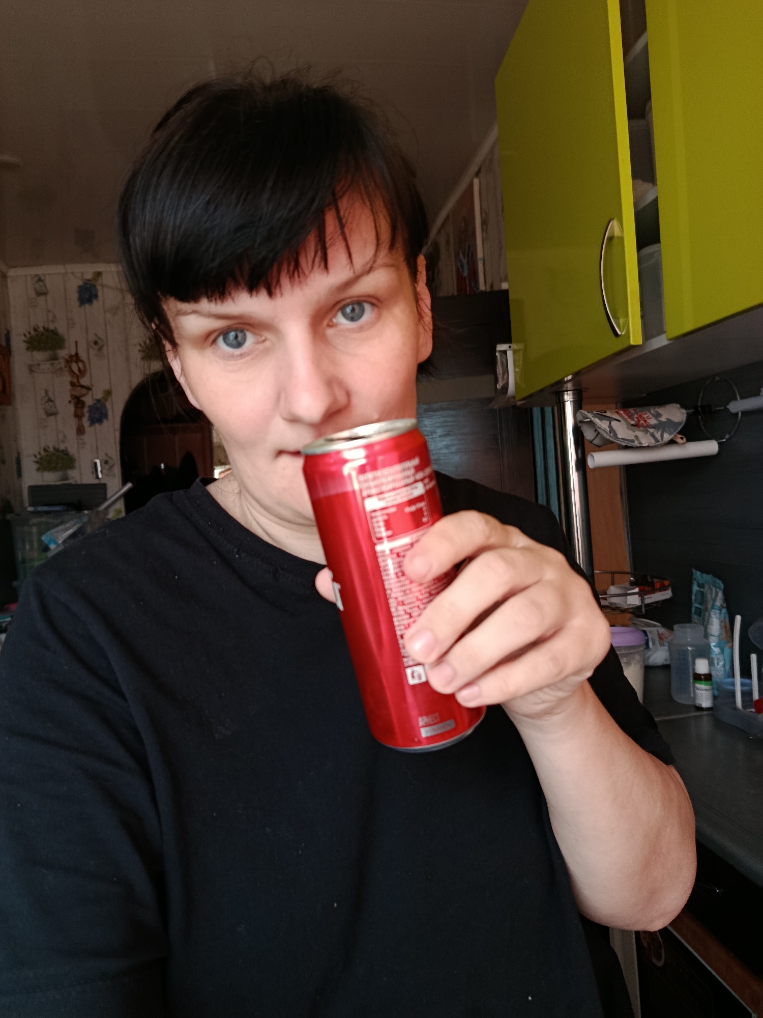 A woman with short, dark hair and blue eyes is captured in a medium shot, looking directly at the camera with a neutral expression. She is wearing a black t-shirt and holds a red aluminum can, likely a soda, near her mouth as if about to drink or having just taken a sip. White text is visible on the can, including "APECT" near the bottom, and other smaller, illegible white text, possibly nutritional information.
The setting appears to be a home kitchen in Фоки, Russia. To the right of the woman, lime green overhead cabinets are prominent, with one door slightly ajar. A dark countertop is visible below the cabinets, adorned with various containers, bottles, and other kitchen items. A silver support pole and a white rail are also part of the kitchen fixtures. The wall behind the woman features patterned wallpaper with a cream background and blue and green floral or leaf designs. An arched doorway is visible in the background on the left, suggesting another room beyond. The lighting suggests an indoor daytime scene.