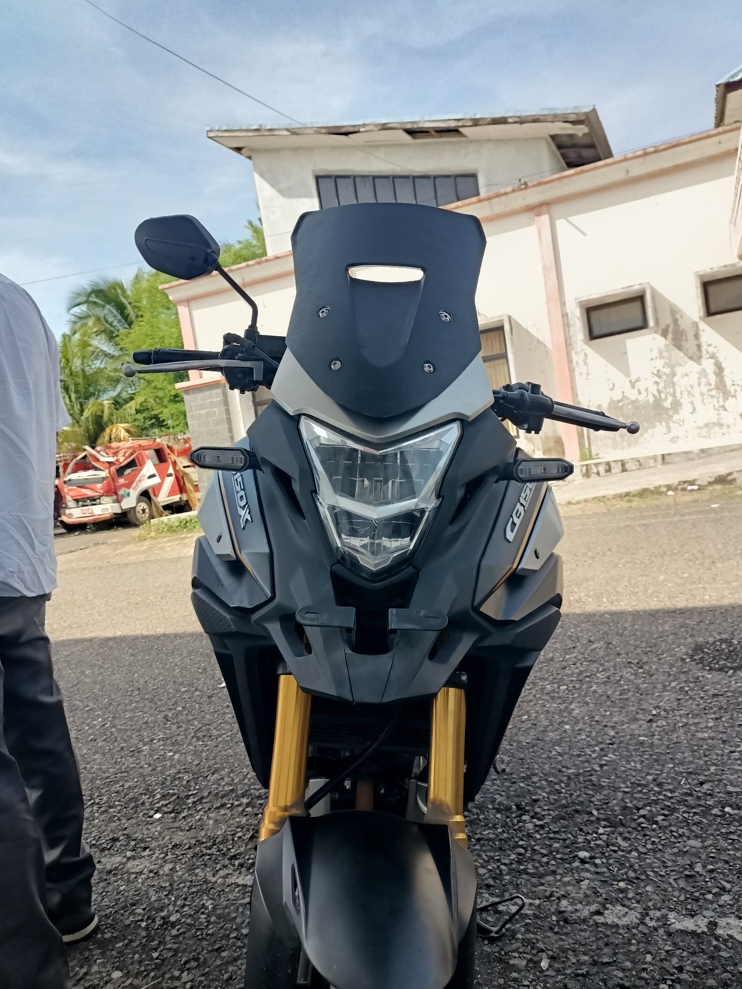 A front-facing, eye-level medium shot captures the front of a black and gray motorcycle with gold front suspension. The motorcycle is parked on a gravel surface in front of a dilapidated white building with an orange trim. In the background, to the left of the motorcycle, a damaged red truck is visible. The sky above is a bright blue with some scattered clouds. A person, partially visible on the left side of the frame, stands facing away from the camera, wearing dark pants and a white shirt. The scene appears to be set in the city of Blangbladeh, Indonesia.