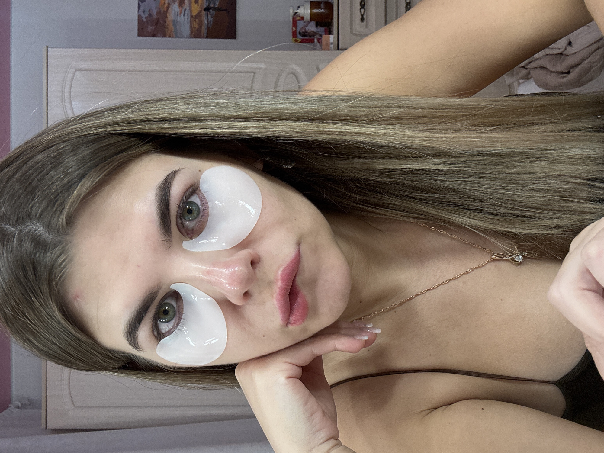 A young adult female is depicted in a close-up, chest-up portrait within an indoor domestic setting. She possesses light skin, long, straight light brown hair with blonde highlights, and green eyes. Two white, translucent, gel-like under-eye patches are affixed beneath her eyes. Her nose exhibits a reddish coloration. Her lips are pink and slightly parted. A small, red blemish is visible on her forehead. Her right hand is positioned with curled fingers, supporting her chin. She wears a dark-colored, thin-strapped garment and a gold-colored chain necklace with a small, clear-stoned pendant. Her fingernails are manicured with clear polish. The background consists of light-colored walls. To the left of the subject, a portion of a light-colored wooden wardrobe with paneled doors is visible. To the right, a light-colored wooden dresser is present, upon which a framed landscape painting, a clear bottle with a pump dispenser containing brown liquid, and a small framed photograph are displayed. A light-colored curtain is partially visible on the far left. The exact location within the city of Chaikovsky, Russia, cannot be determined from the visual evidence in the image.