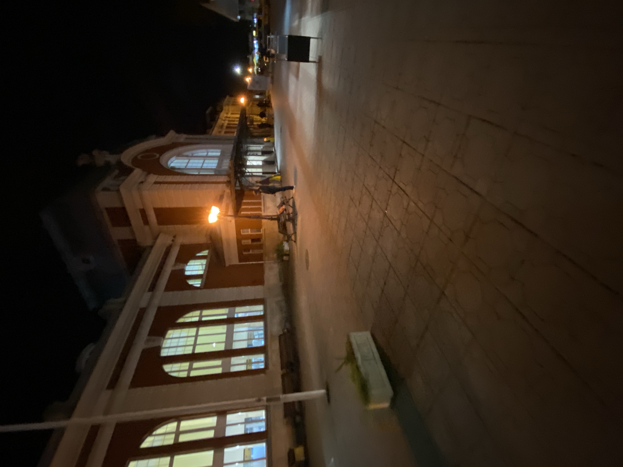 The image shows the Varna Central railway station, Bulgaria at night. The building is brick with white trim. Large arched windows are visible. The foreground shows the pavement with street lights. People are standing on the pavement near the building.