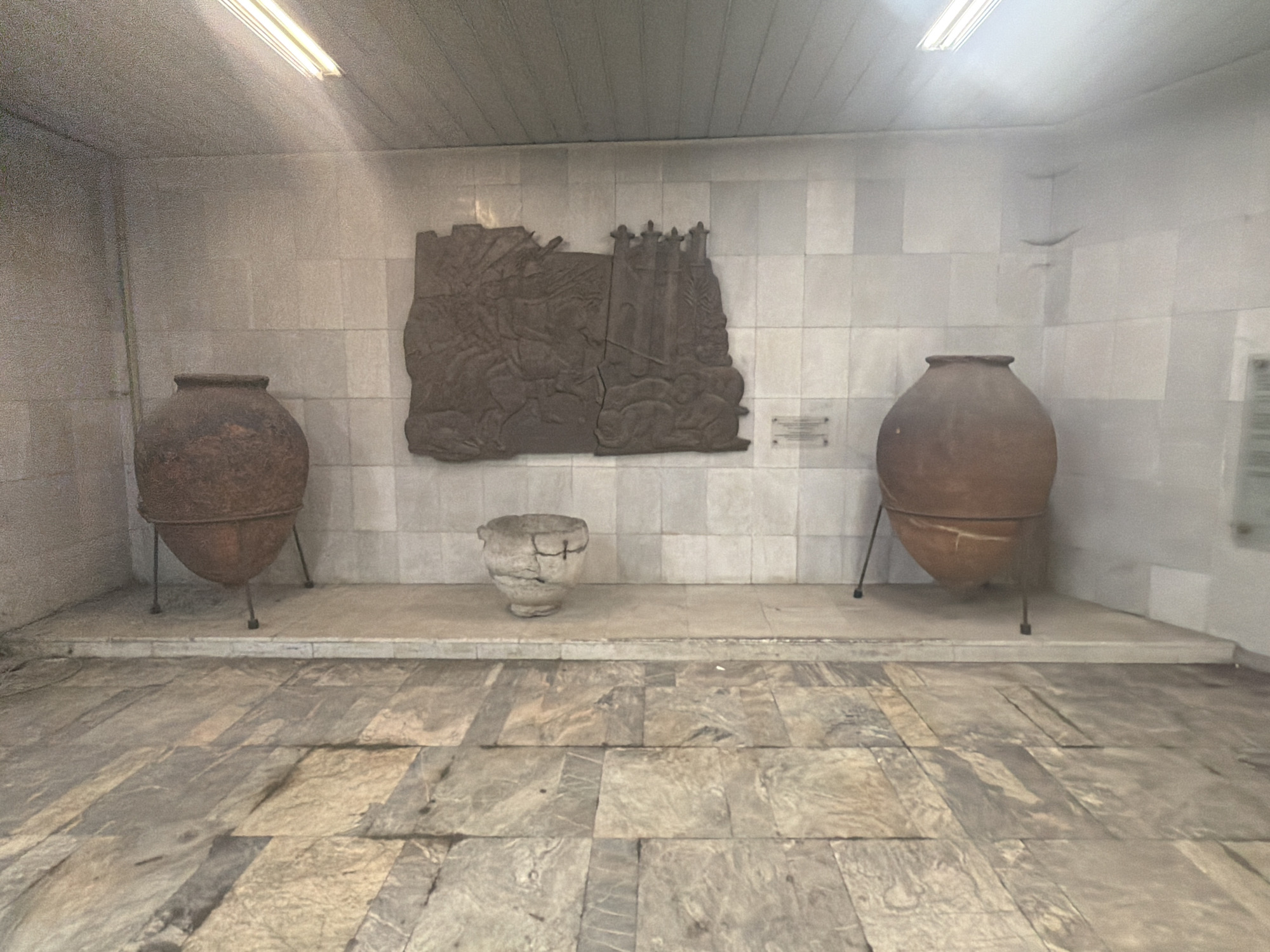 This is an indoor shot of a museum exhibit, likely in Sofia, Bulgaria. The exhibit features three ceramic vessels - two large amphora-like pots on stands, flanking a smaller bowl-shaped vessel. Behind them is a large bas-relief sculpture depicting a historical scene with figures and possibly structures. The backdrop is a wall tiled with large, light-colored rectangular tiles. The floor is paved with large, irregularly shaped stone tiles. The lighting is somewhat dim, with some overhead artificial lights visible. There are small informational plaques on the wall next to the relief and on the right-hand side of the shot. There are no people visible in the frame. The general atmosphere suggests a historical or archaeological museum setting.