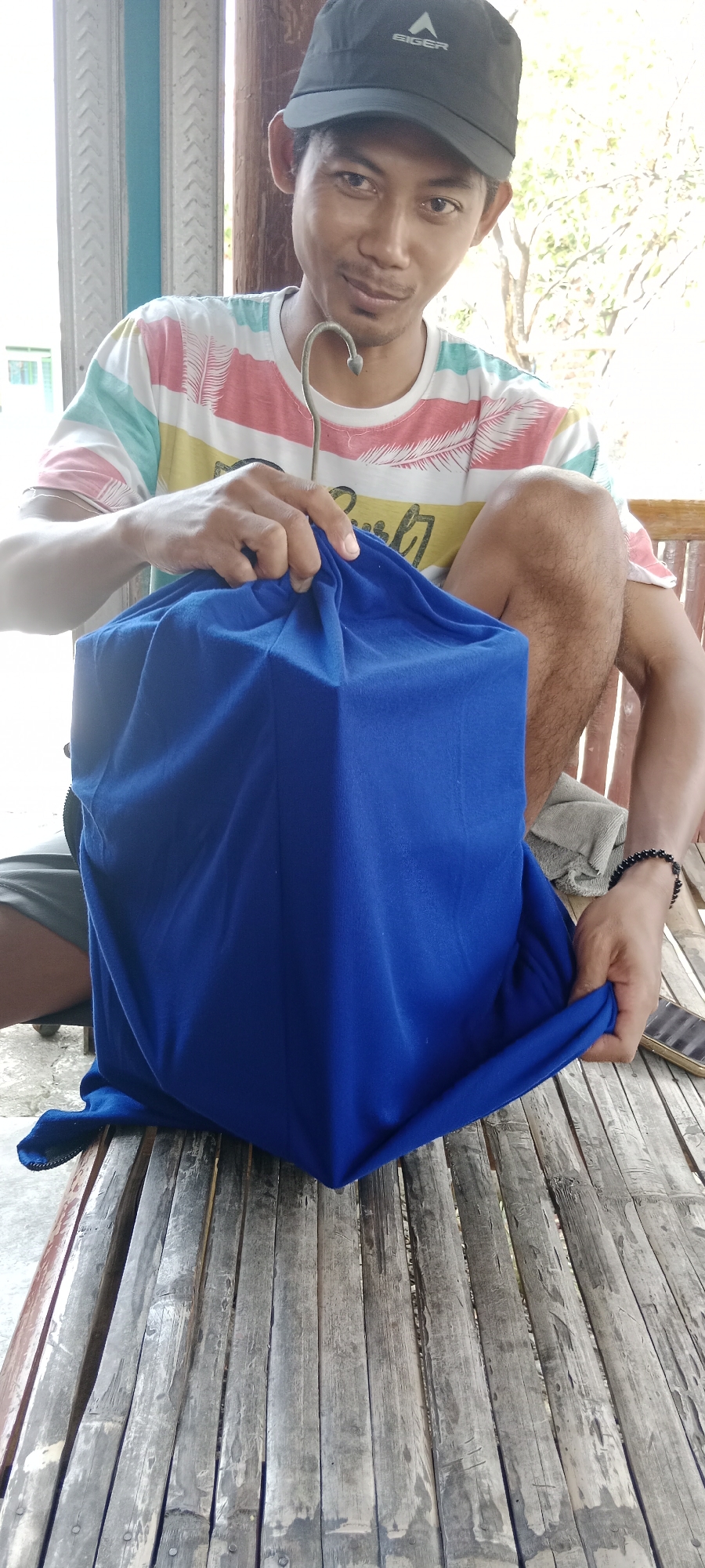 A man wearing a black baseball cap and a colorful striped t-shirt is sitting on a bamboo bench outdoors. He is holding a large, cubic object draped in a bright blue cloth. The man's hands are visible as he adjusts the cloth. A metal hook is attached to the object and is held by the man. In the background, there are wooden pillars and foliage, suggesting an outdoor setting. The bench is made of aged, weathered bamboo slats. A mobile phone and a dark beaded bracelet are visible on the man's wrist. The overall impression is a casual, informal scene.