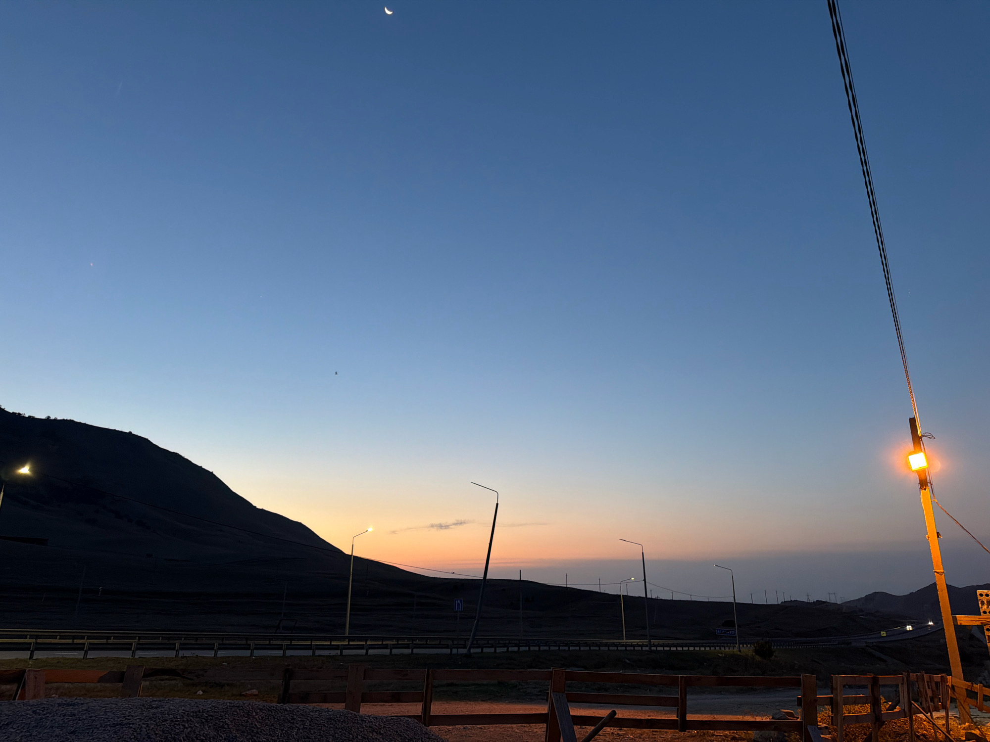 This is a photograph taken at dusk in Agachaul, Russia. The sky is a gradient of deep blue at the top, transitioning to lighter blues and a faint orange and pink near the horizon. A thin crescent moon is visible in the upper left portion of the sky. A mountain range forms the dark silhouette on the left side of the image. A road with streetlights runs horizontally across the middle ground. Several streetlights are visible, some illuminated and casting a warm glow. Power lines are strung diagonally across the right side of the image, with one pole featuring a bright, square-shaped yellow light. In the foreground, a wooden fence and piles of gravel are visible. The overall scene depicts a rural or roadside landscape at twilight.