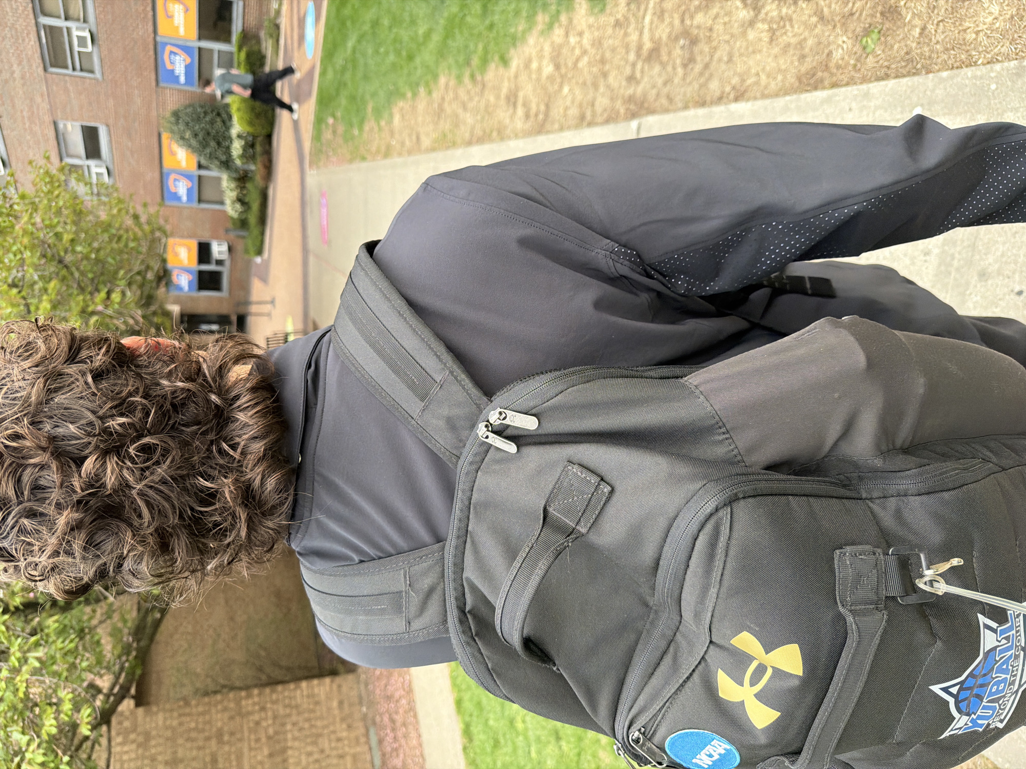 A waist-level shot looking down shows a person wearing a gray shirt and gray pants, with a black Under Armour backpack. The backpack has a gold logo, a "Basketball Beyond the Court" patch, and an NCAA patch. The person has curly brown hair. They are standing on a sidewalk next to a grassy area. In the background, there is a brick building with blue and orange signs. A person in a green shirt and black pants is walking on the sidewalk away from the viewer.