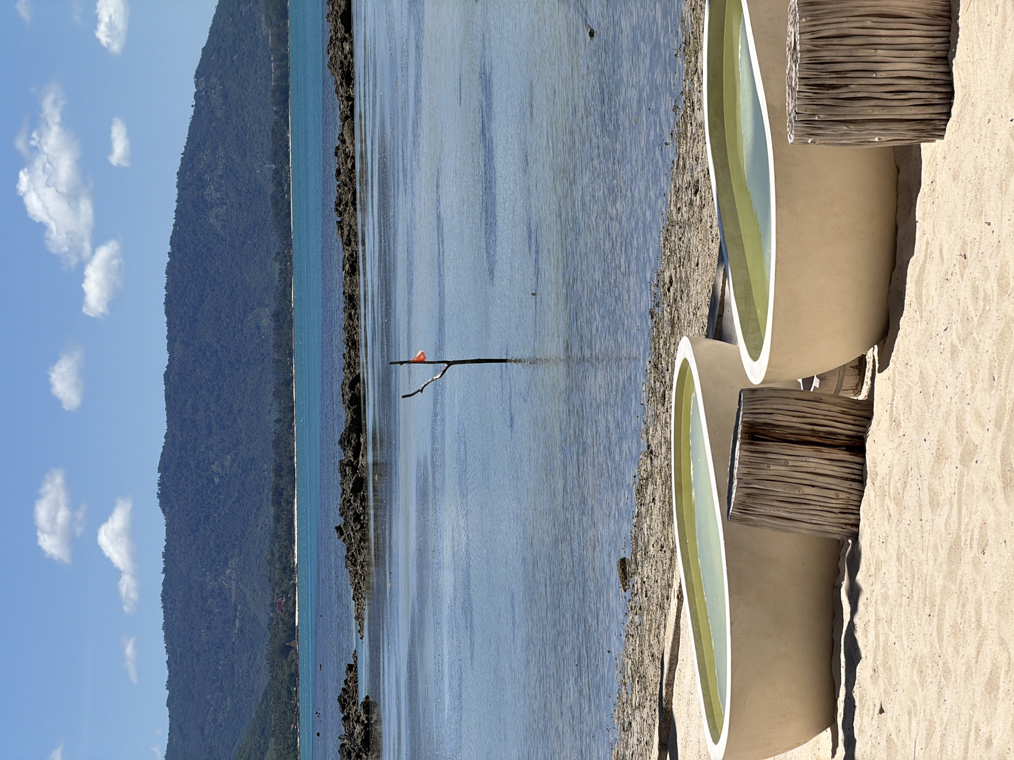This is a vertical, eye-level, medium shot of a serene beach scene in Baan Thong Krut, Thailand. The foreground features two beige, crescent-shaped bathtubs with light green water, placed on a sandy shore. A rustic, cylindrical wooden structure stands between them, adding a natural element to the setting.
In the midground, the calm, blue water gently laps against the shore, with a line of dark, rugged rocks separating the beach from the deeper sea. A single, thin branch extends from the water, with a small, bright orange object, possibly a piece of plastic or fabric, tied to it. The water's surface reflects the bright blue sky and scattered white clouds.
The background is dominated by a lush, green, tree-covered mountain rising from the water's edge. The sky is a clear, vibrant blue, with a few fluffy white clouds drifting by. The overall atmosphere is peaceful and tranquil, suggesting a warm, sunny day, likely mid-morning or early afternoon, given the bright lighting and clear skies. There is no visible text in the image.